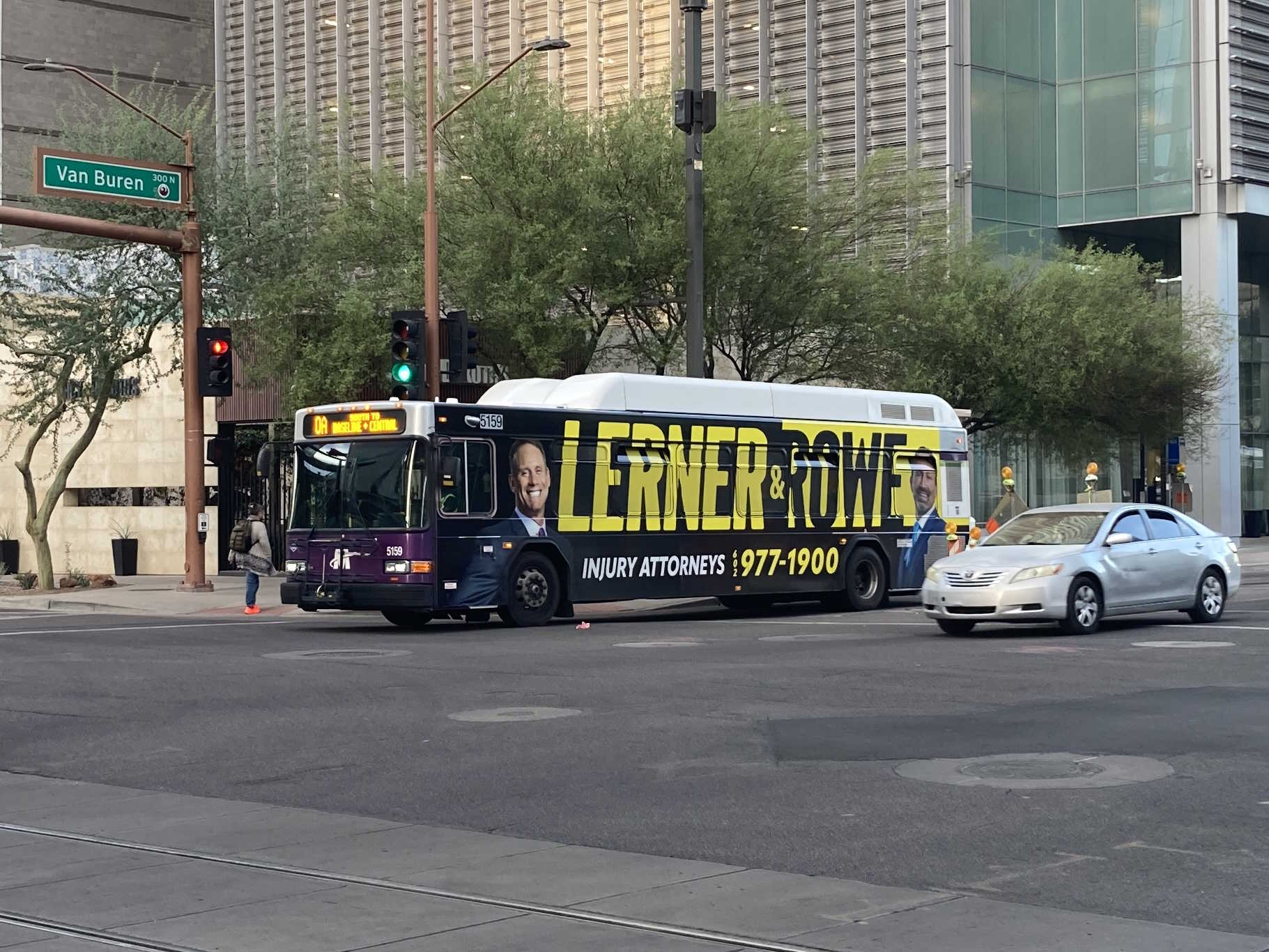 A white and gray Valley Metro bus, with purple and green accent colors, number 5159, traveling westbound on Van Buren Street in Phoenix on route 0A to Baseline and Central