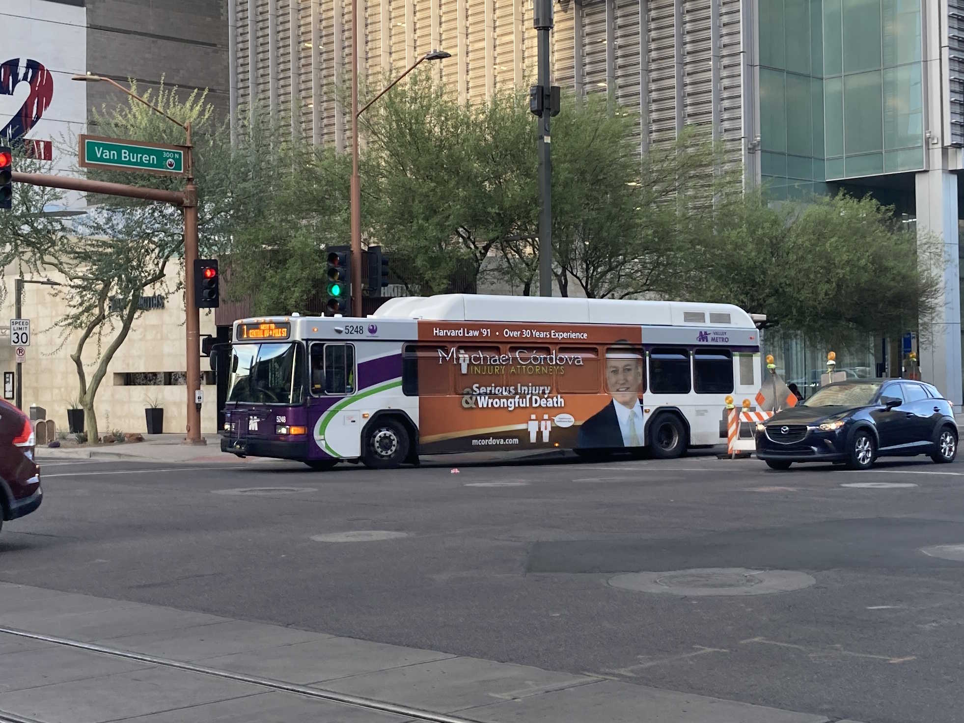 A white and gray Valley Metro bus, with purple and green accent colors, number 5248, traveling westbound on Van Buren Street in Phoenix on route 1 to Central Avenue and Polk Street