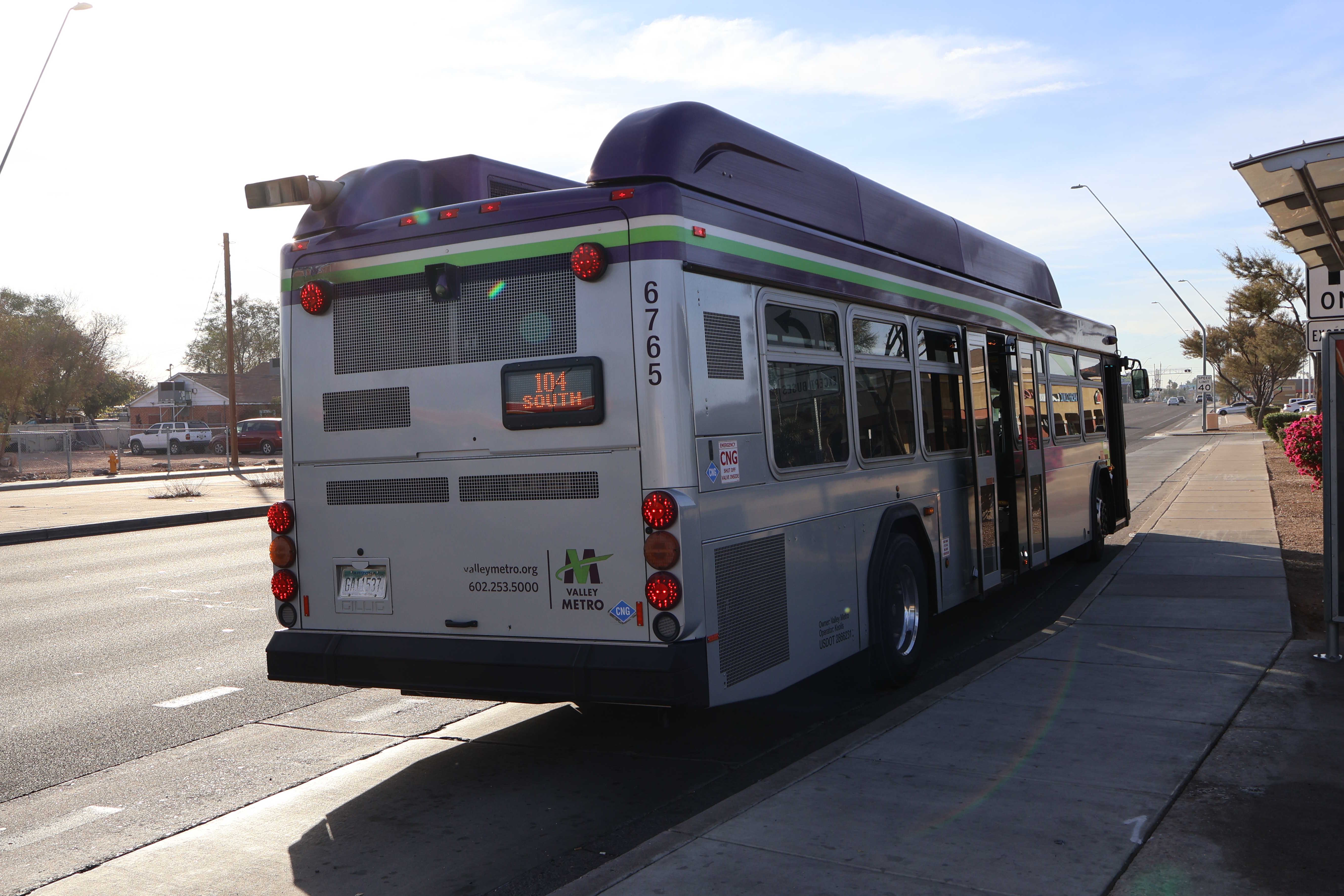 A gray and purple Valley Metro bus with a white and green stripe, number 6765, traveling southbound on Alma School Road in Mesa on route 104 to Morelos Street and Hamilton Street