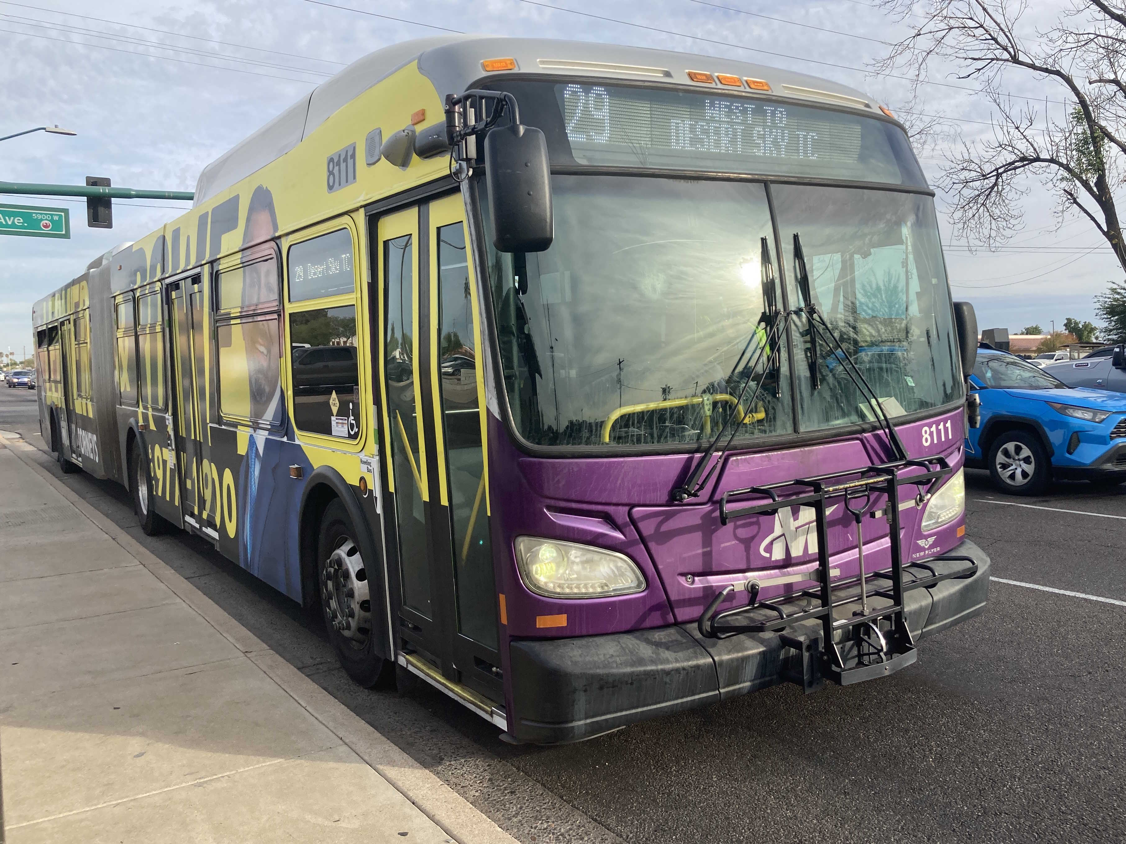 A white and gray articulated Valley Metro bus, with purple and green accent colors, number 8111, traveling westbound on Thomas Road in Phoenix on route 29 to Desert Sky Transit Center