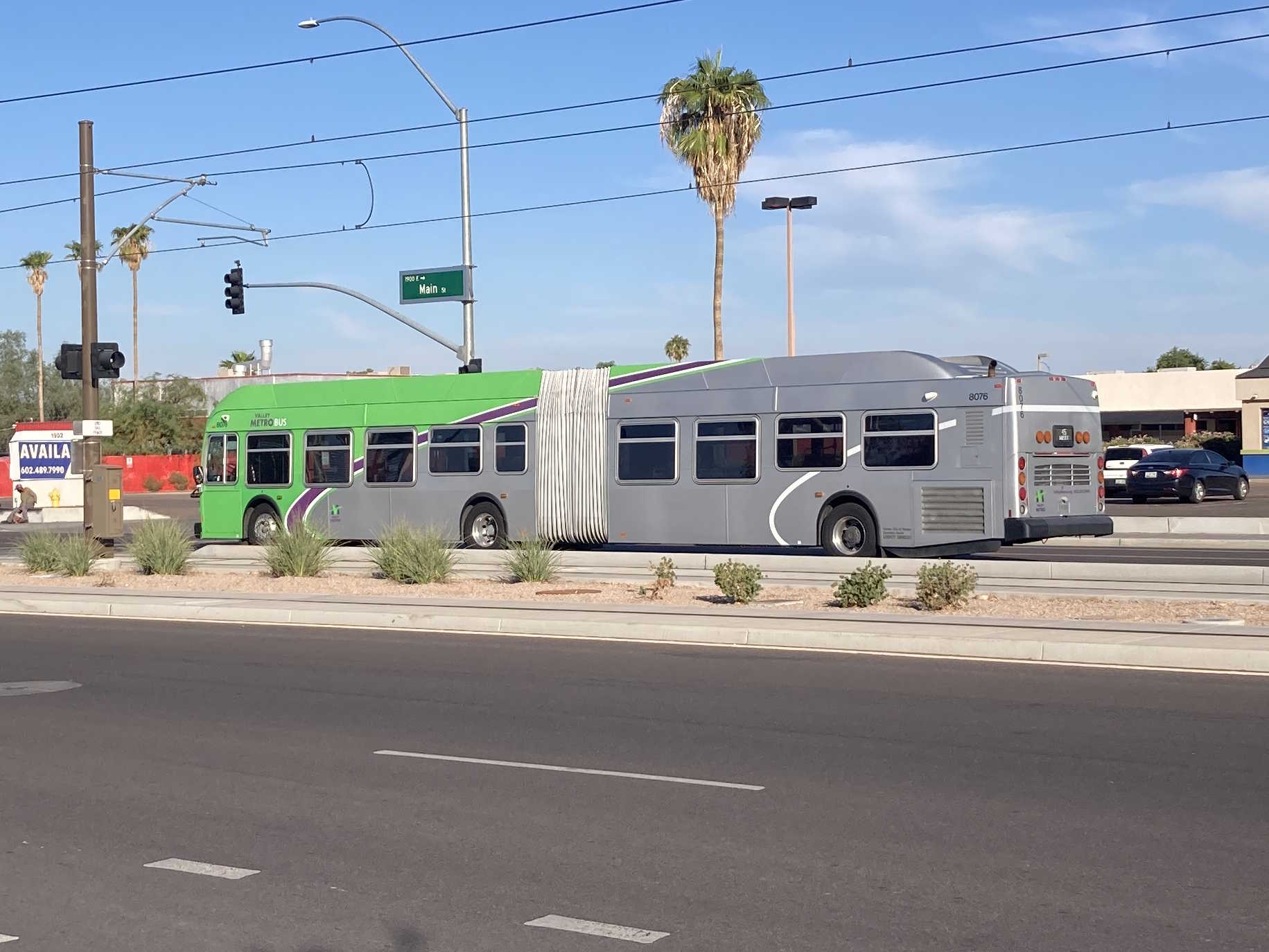 A gray and green articulated Valley Metro bus with a purple and white stripe, number 8076, traveling westbound on Main Street in Mesa on route 45 to Broadway Road and 19th Avenue