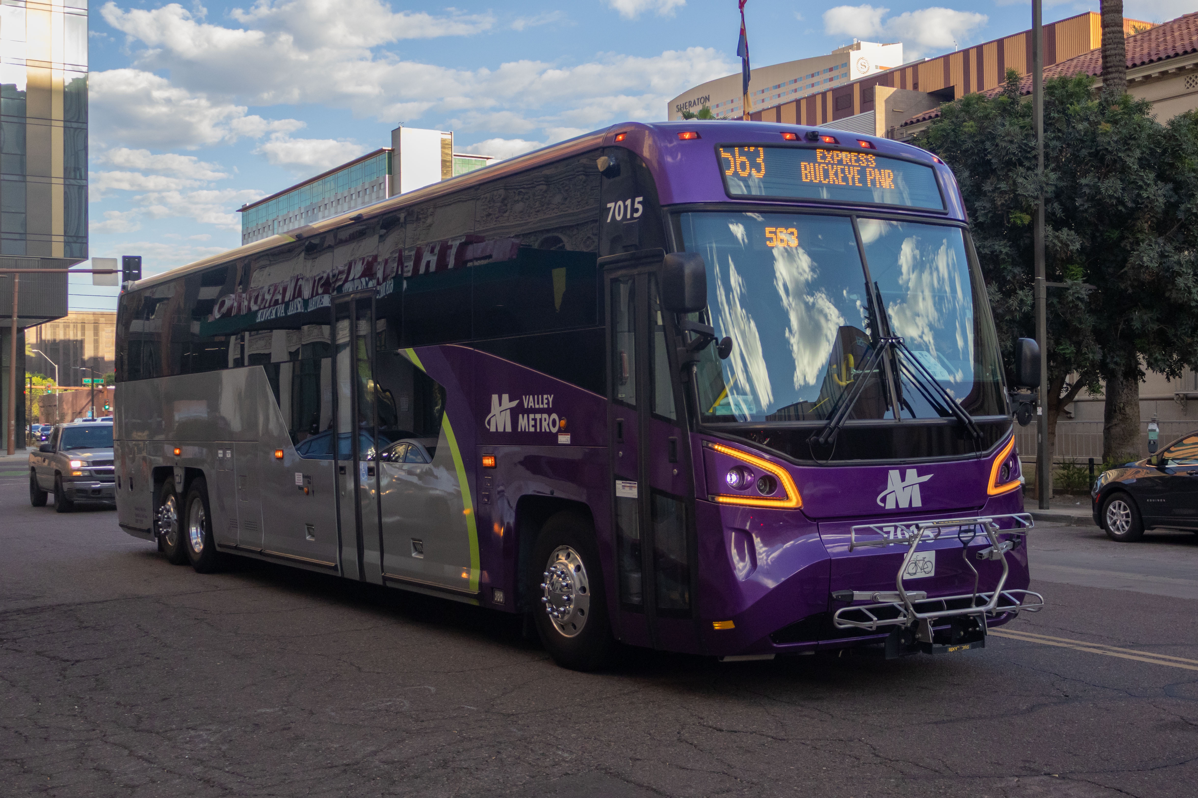 A gray and purple Valley Metro coach bus, with a green stripe, number 7015, traveling westbound on Fillmore Street in Phoenix on route 563 to Buckeye Park and Ride