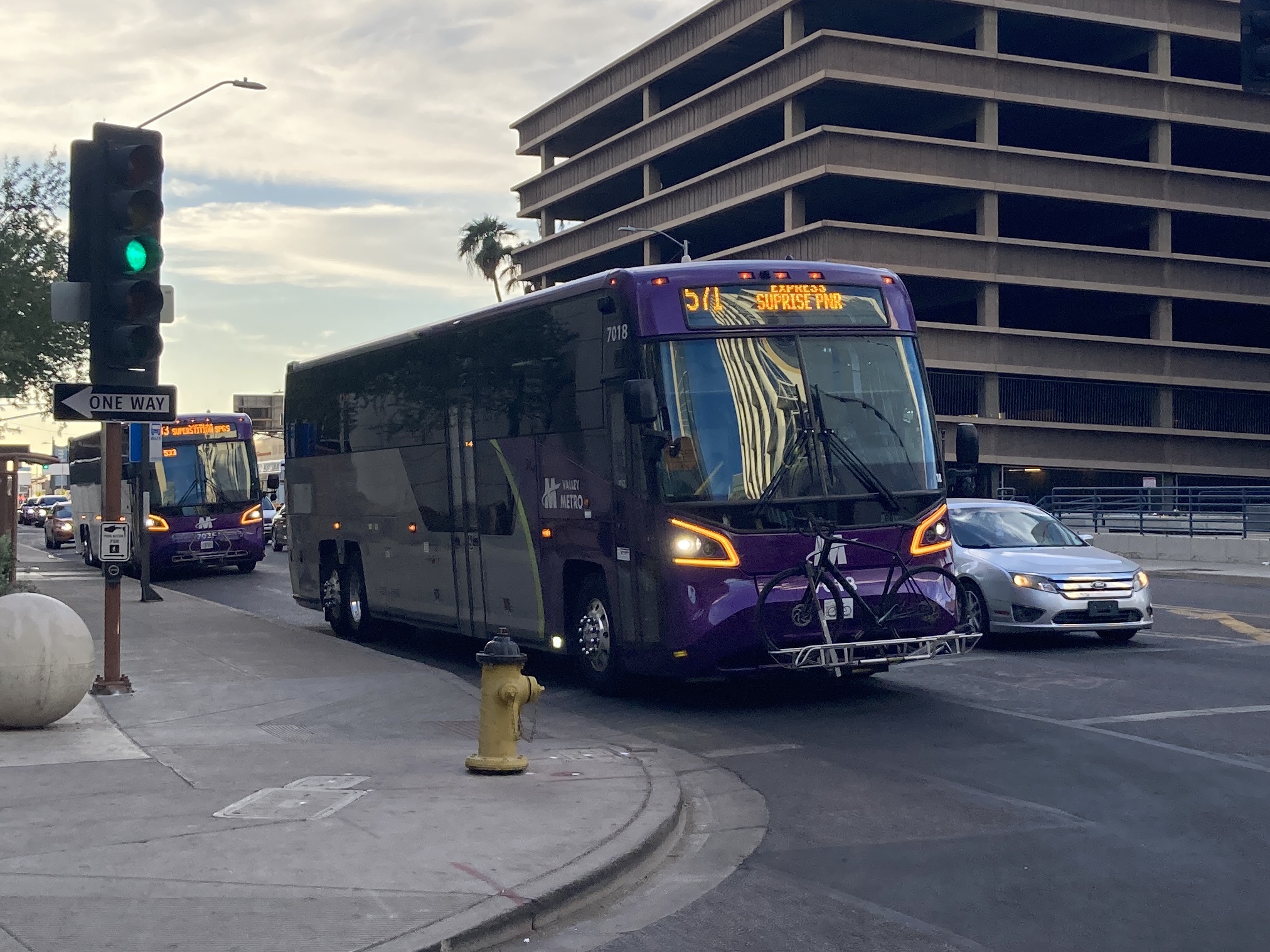 A gray and purple Valley Metro bus, with a green stripe, number 7018, traveling eastbound on Van Buren Street in Phoenix on route 571 to Surprise Park and Ride