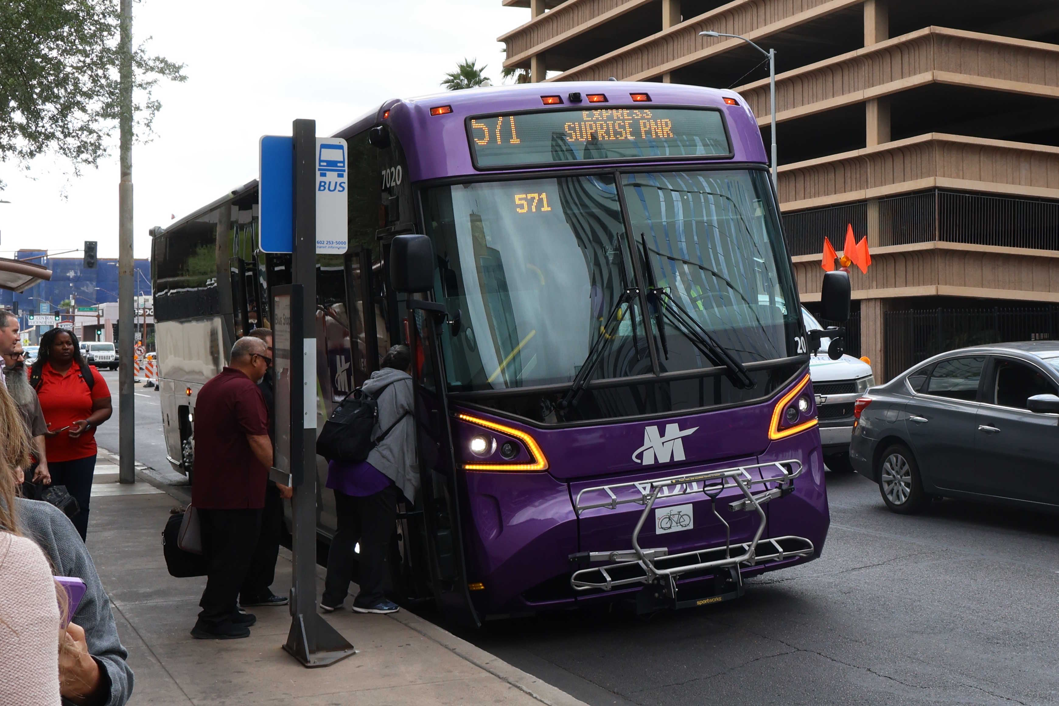 A gray and purple Valley coach Metro bus, with a green stripe, number 7020, traveling eastbound on Van Buren Street in Phoenix on route 571 to Surprise Park and Ride