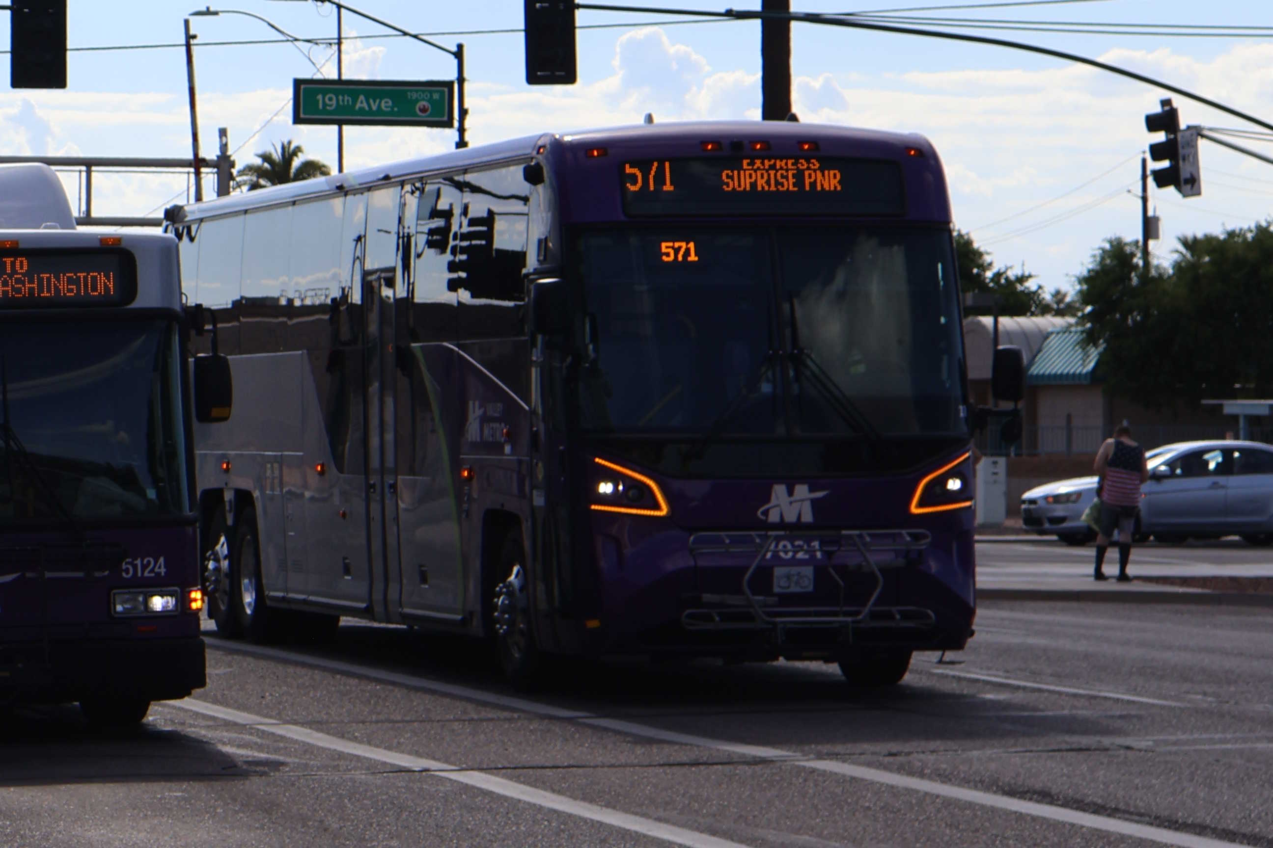 A gray and purple Valley Metro coach bus, with a green stripe, number 7021, traveling eastbound on Jefferson Street in Phoenix on route 571 to Surprise Park and Ride