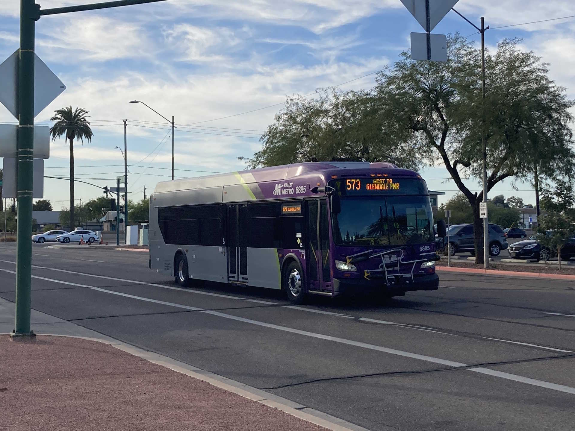 A gray and purple Valley Metro bus, with a green stripe, number 6885, traveling eastbound on Jefferson Street in Phoenix on route 573 to Glendale Park and Ride