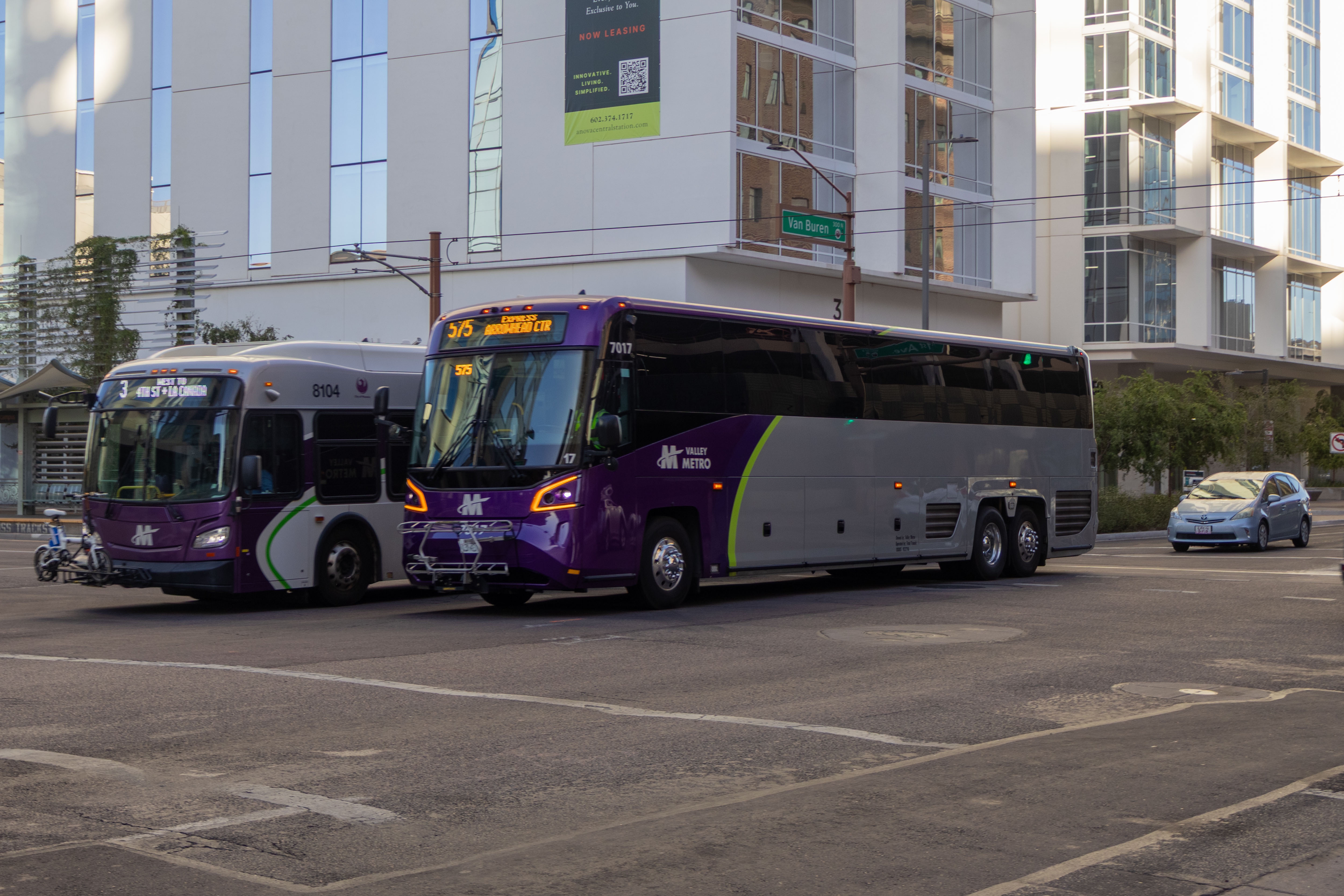 A gray and purple Valley Metro coach bus, with a green stripe, number 7017, traveling westbound on Van Buren Street in Phoenix on route 575 to Arrowhead Mall Transit Center