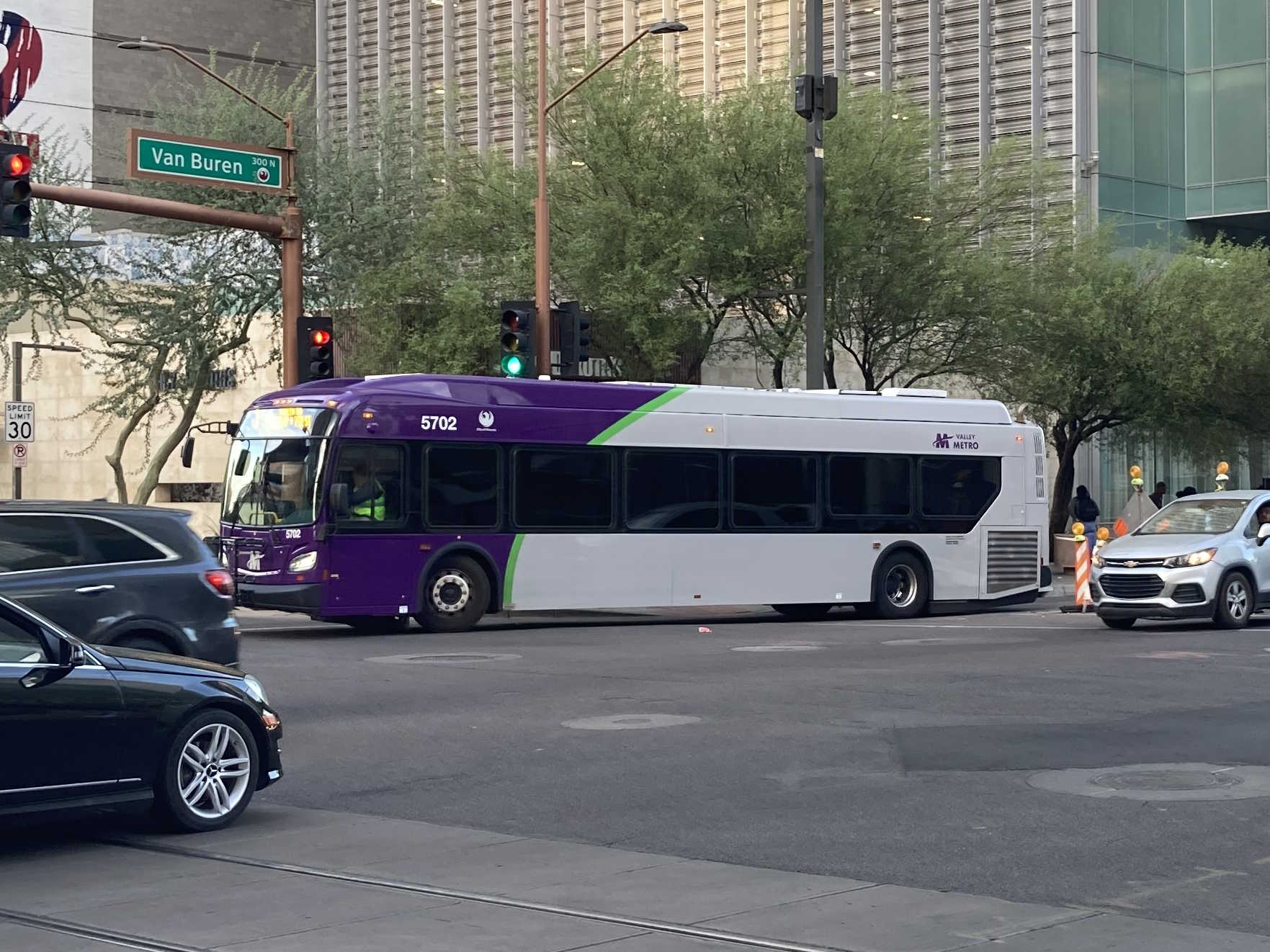 A gray and purple Valley Metro bus, with a green stripe, number 5702, traveling westbound on Van Buren Street in Phoenix on route 7 to 7th Street and Dunlap Avenue