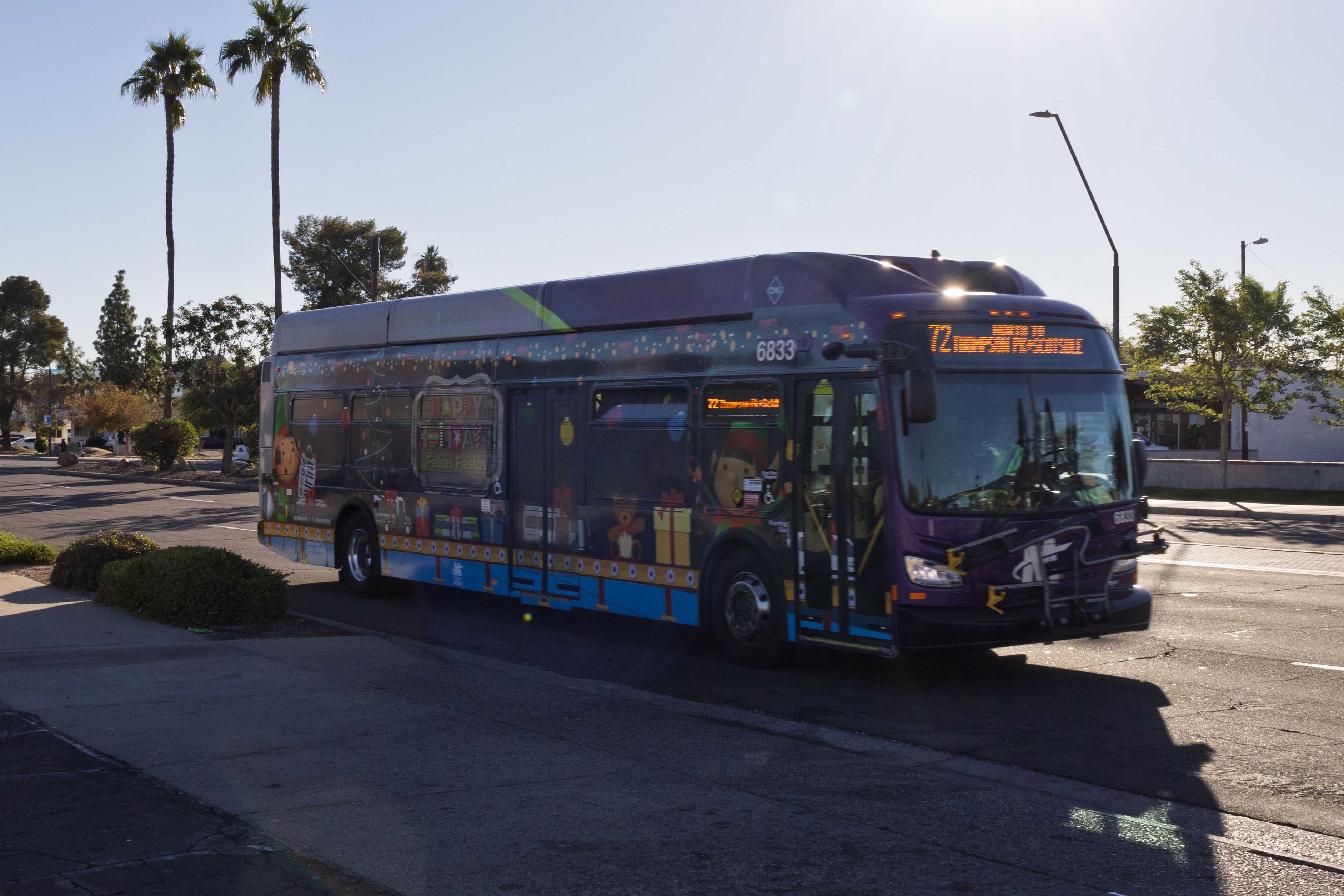 A gray and purple Valley Metro bus, with a green stripe and a Happy Holidays wrap, number 6833, traveling northbound on Scottsdale Road in Tempe on route 72 to Thompson Peak Parkway and Scottsdale Healthcare