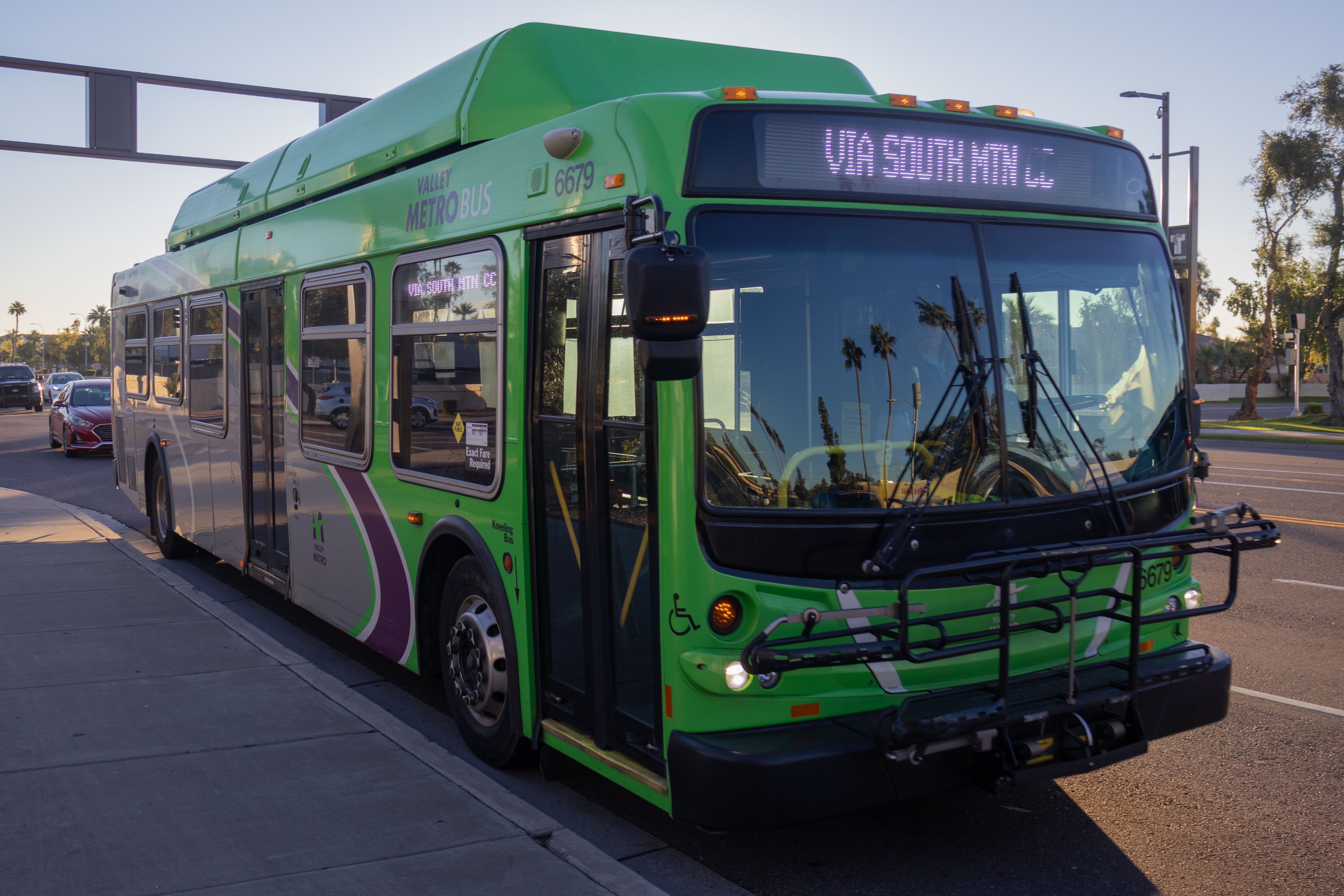 A gray and green Valley Metro bus, number 6679, traveling westbound on Baseline Road in Tempe on route 77 to Gila River Service Center via South Mountain Community College