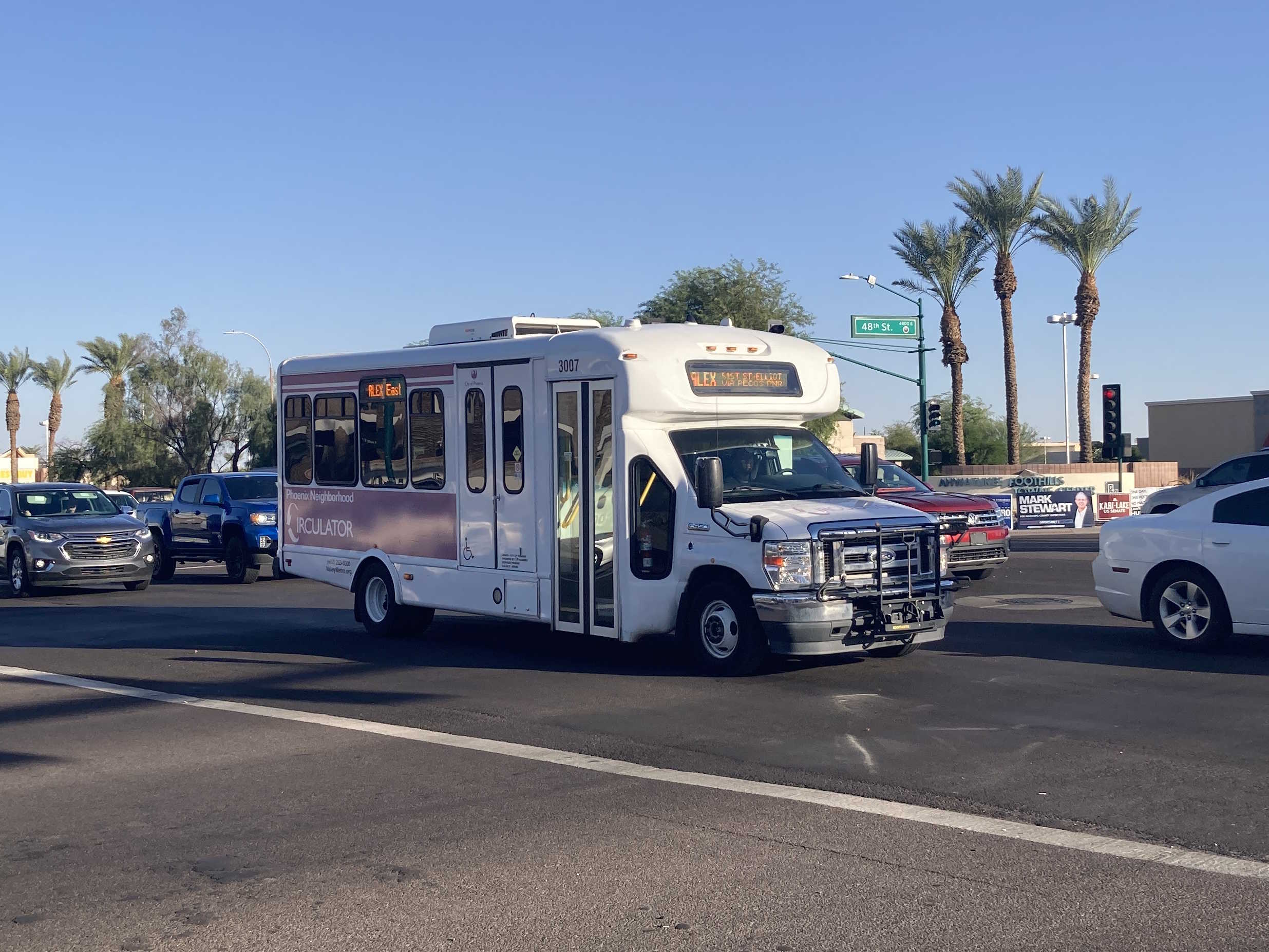 A white and purple Phoenix Neighborhood Circulator minibus, number 3007, traveling westbound on Ray Road in Phoenix on the ALEX route to 51st Street and Elliot Road