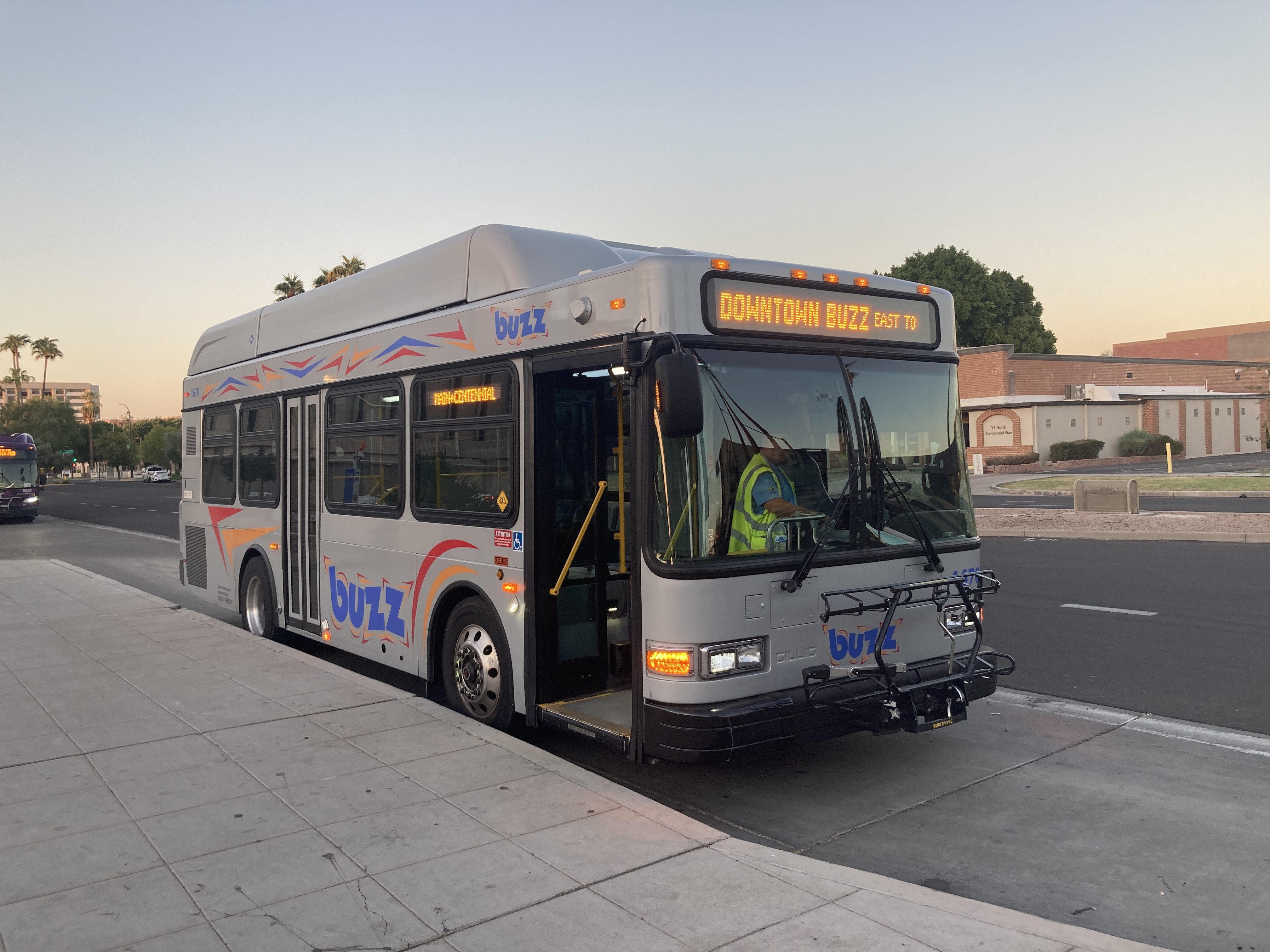 A silver BUZZ bus, with red, orange, and blue accents, number 1670, at Centennial Way and Main Street in Mesa on the Downtown BUZZ route to Centennial Way and Main Street