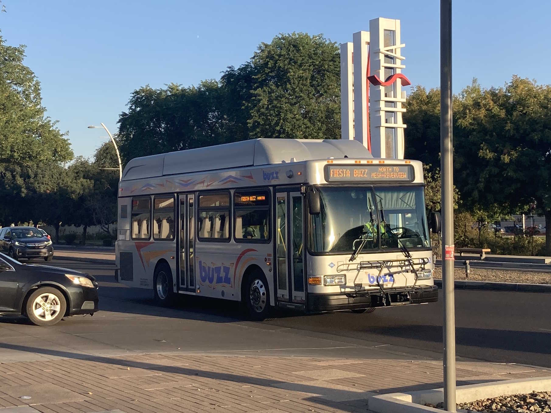 A silver BUZZ bus, with red, orange, and blue accents, number 1672, traveling westbound on Southern Avenue in Mesa on the Fiesta BUZZ route to Mesa Riverview