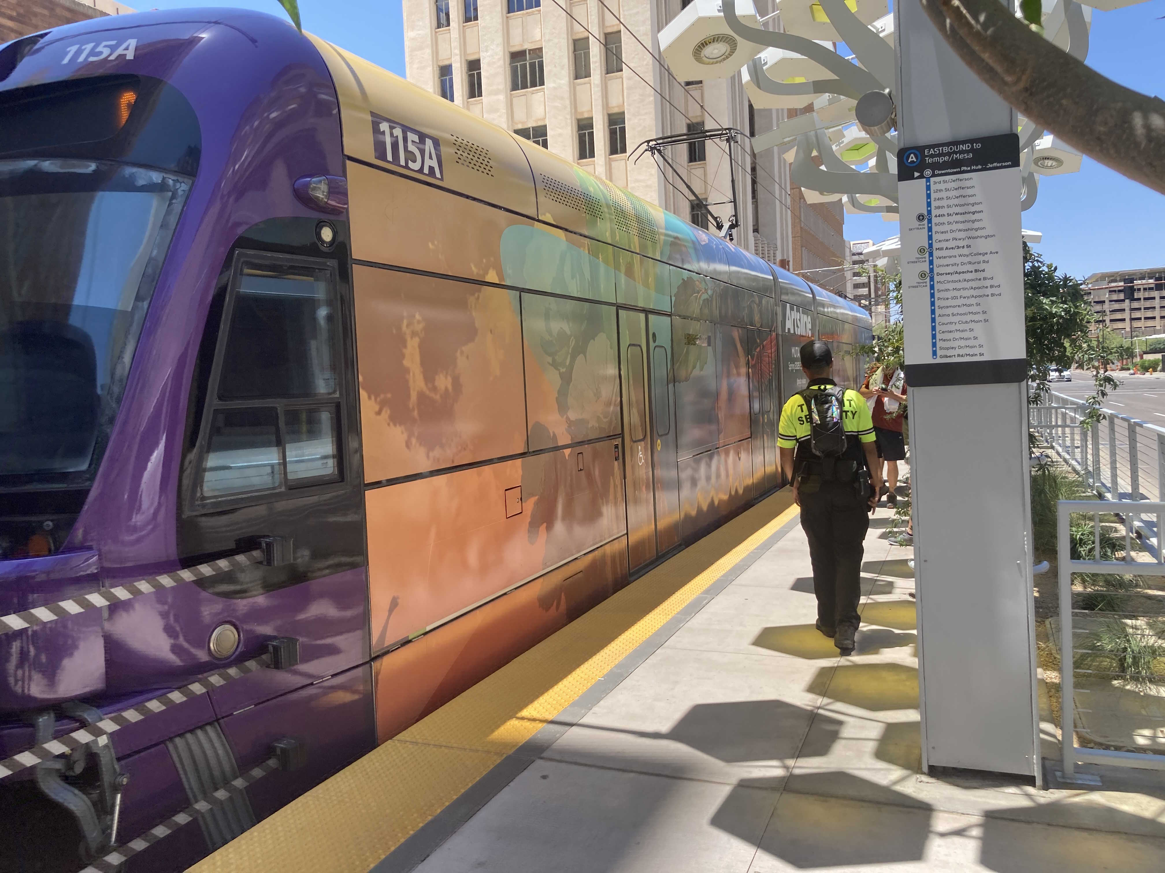 A gray and purple light rail train with a green stripe, number 115, traveling eastbound on Jefferson Street in Phoenix
