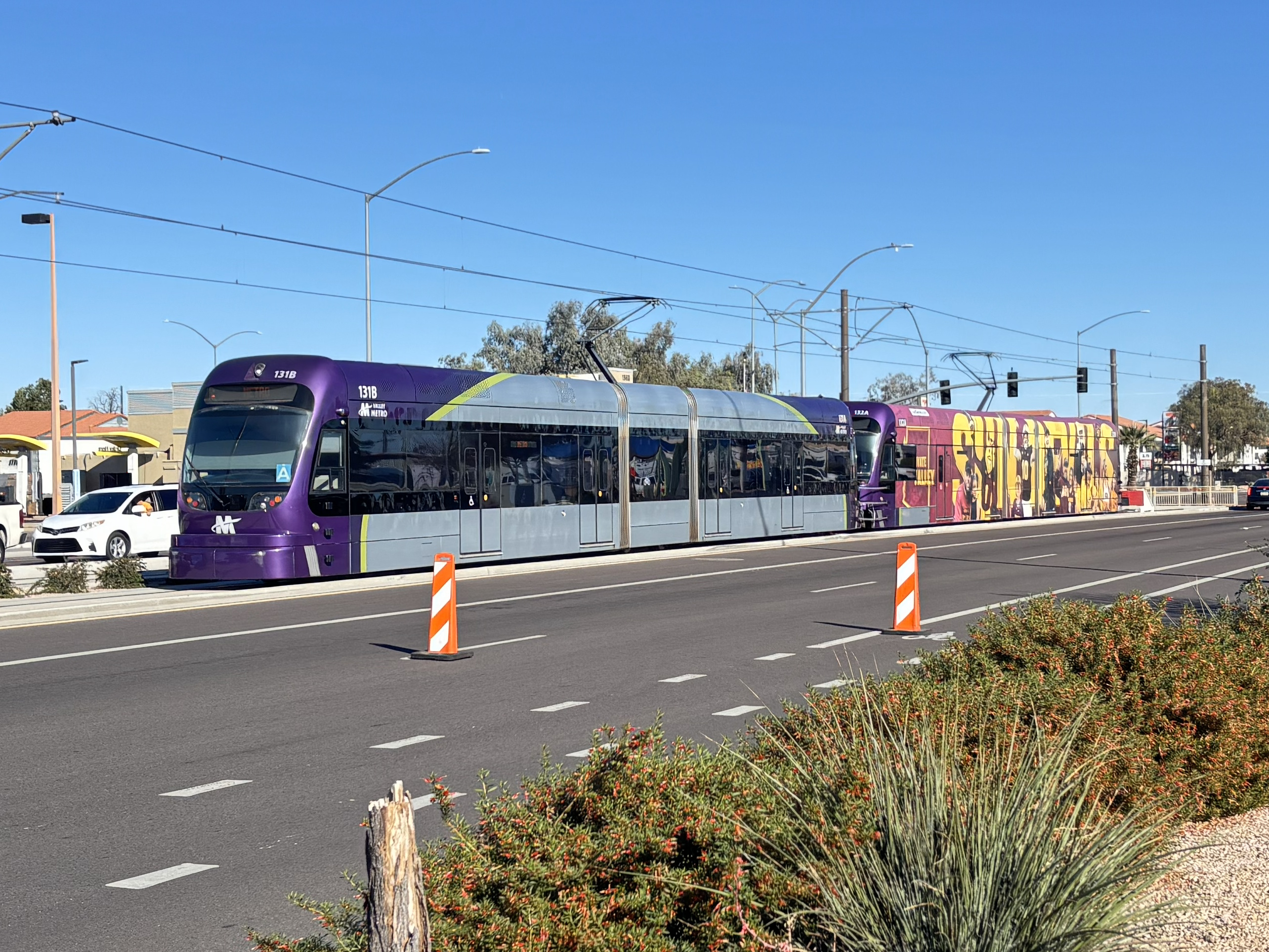 Two gray and purple light rail trains with green stripes, numbers 131 and 132, parked past Gilbert Road and Main Street station in Mesa