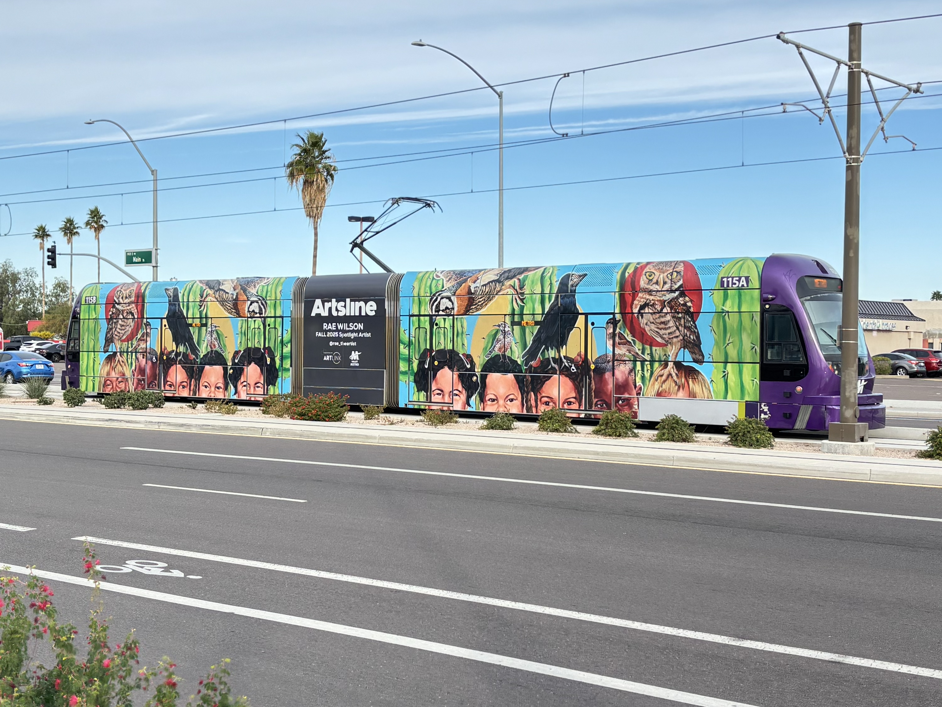 A gray and purple light rail train with a green stripe, number 115, parked past Gilbert Road and Main Street station in Mesa