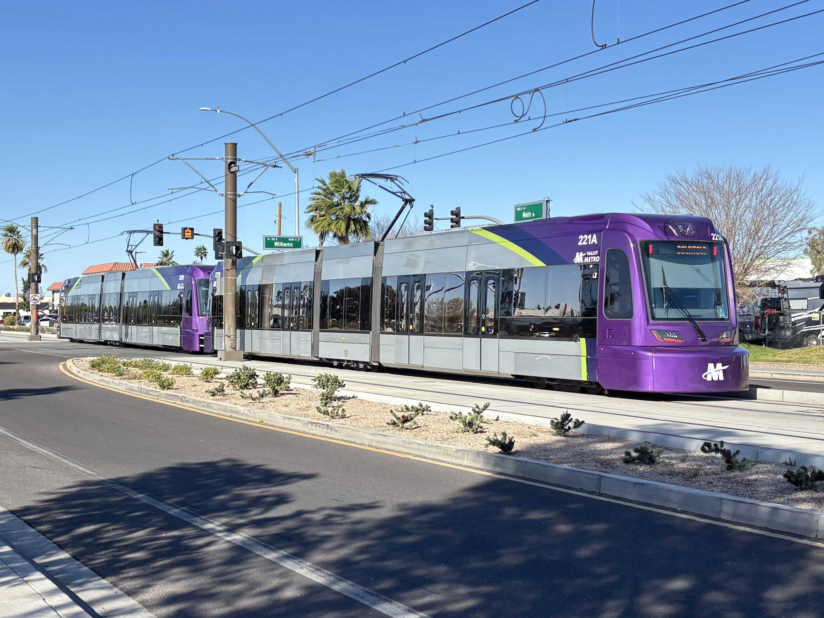 Two gray and purple light rail trains with green stripes, numbers 221 and 222, traveling westbound on Main Street in Mesa