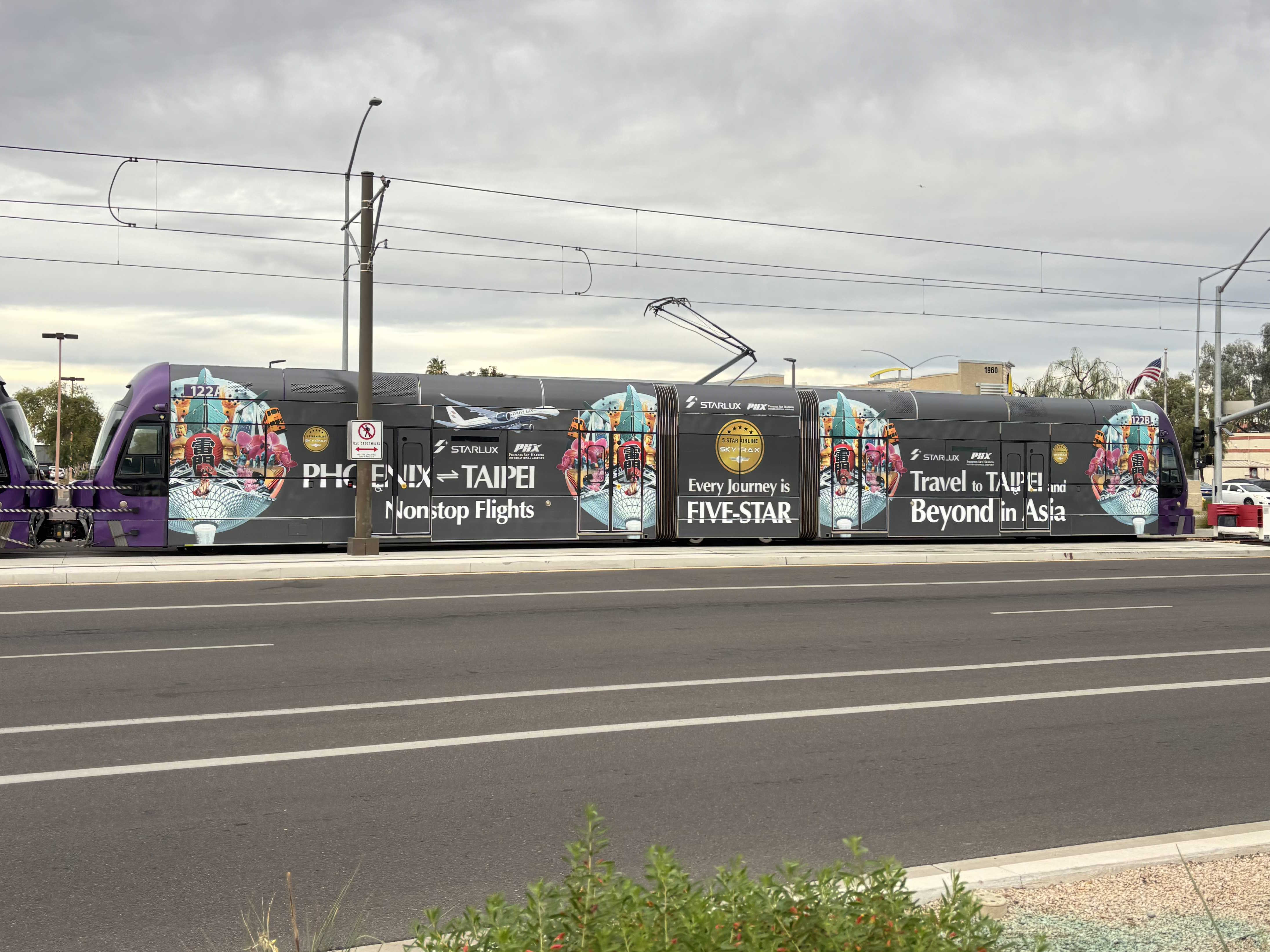 A gray and purple light rail train with a green stripe, number 122, parked past Gilbert Road and Main Street station in Mesa