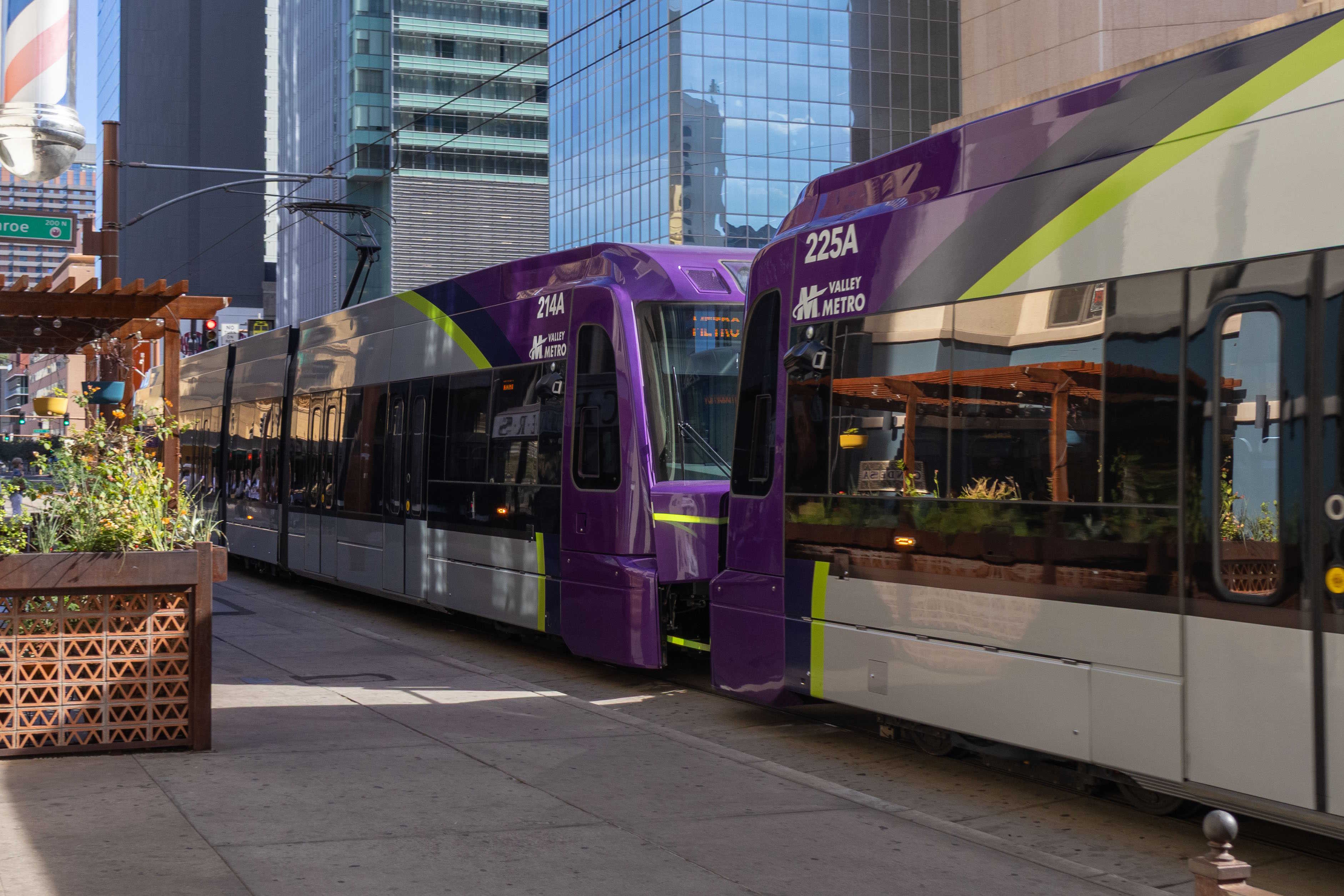 Two gray and purple light rail trains with green stripes, numbers 214 and 225, traveling northbound on Central Avenue in Phoenix