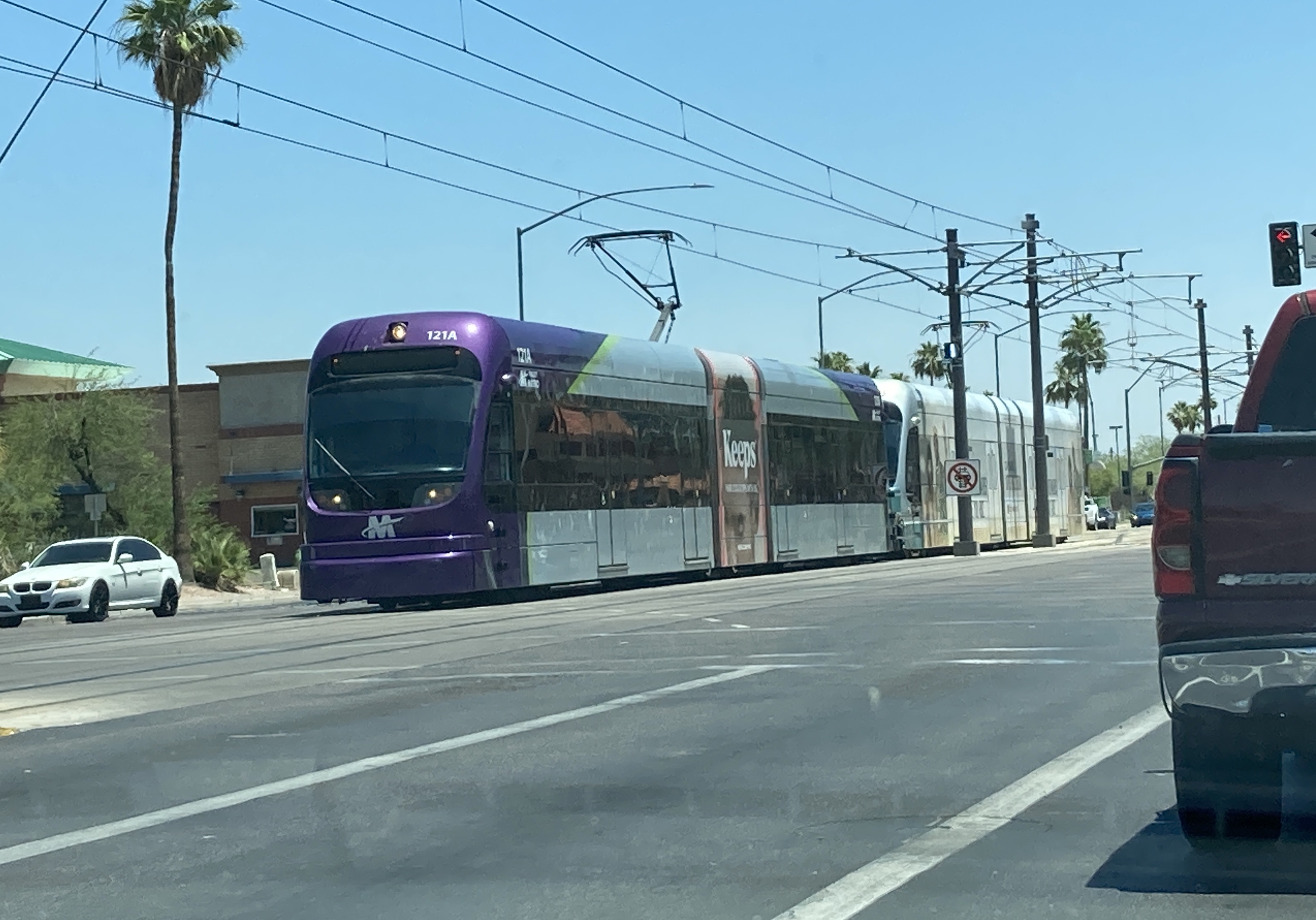 A gray and purple light rail train with a green stripe, number 121, and a silver light rail train, unknown number, traveling westbound on Main Street in Mesa towards 19th Avenue and Dunlap station