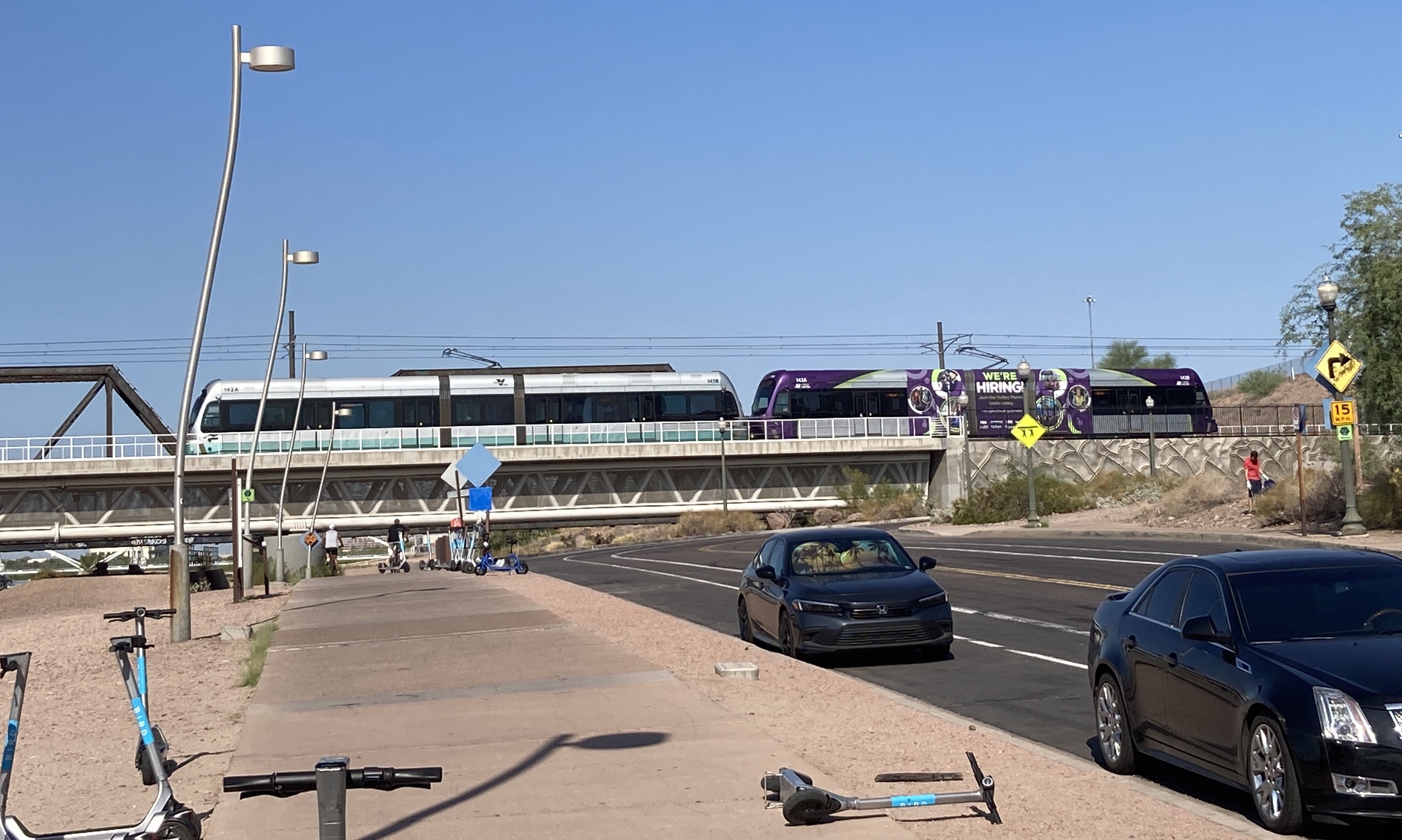 A gray and purple light rail train with a green stripe, number 143, and a silver light rail train, number 142, traveling westbound over Tempe Town Lake towards 19th Avenue and Dunlap station