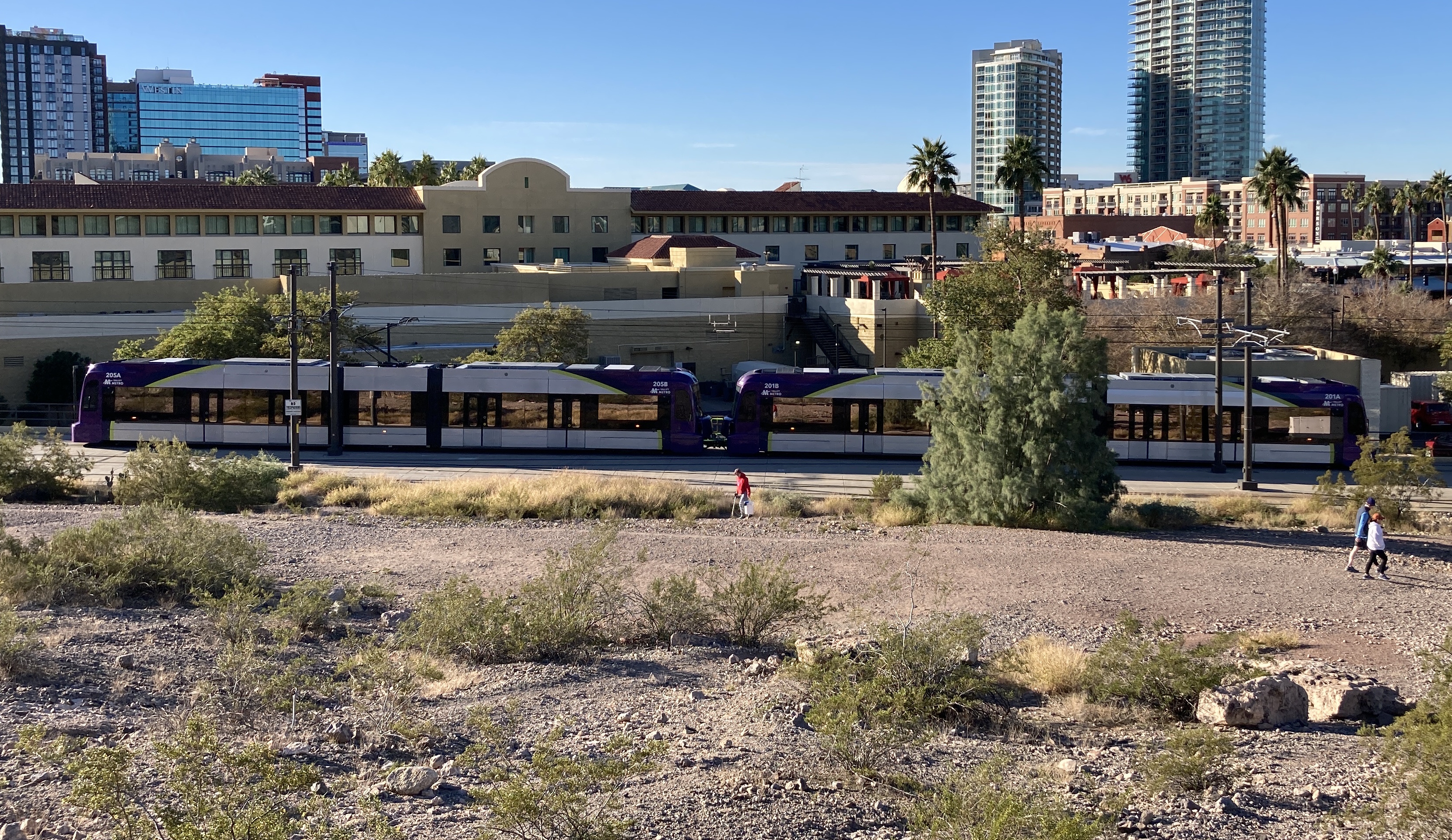 Two gray and purple light rail trains with green stripes, numbers 201 and 205, traveling westbound near 3rd Street and Mill Avenue towards 19th Avenue and Dunlap station