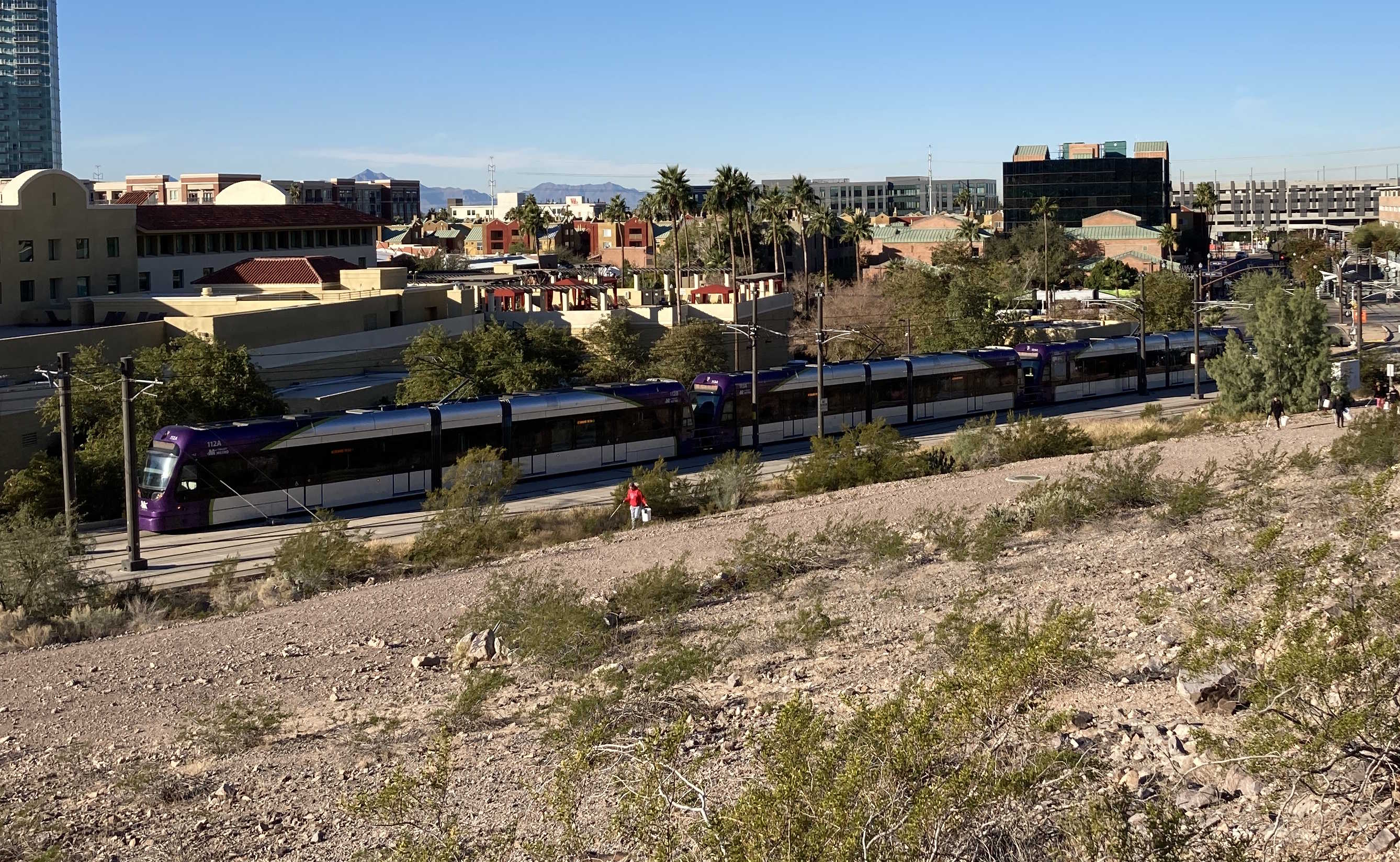 Three gray and purple light rail trains with green stripes, numbers 112, 113, and 125, traveling eastbound near 3rd Street and Mill Avenue towards Gilbert Road and Main Street station