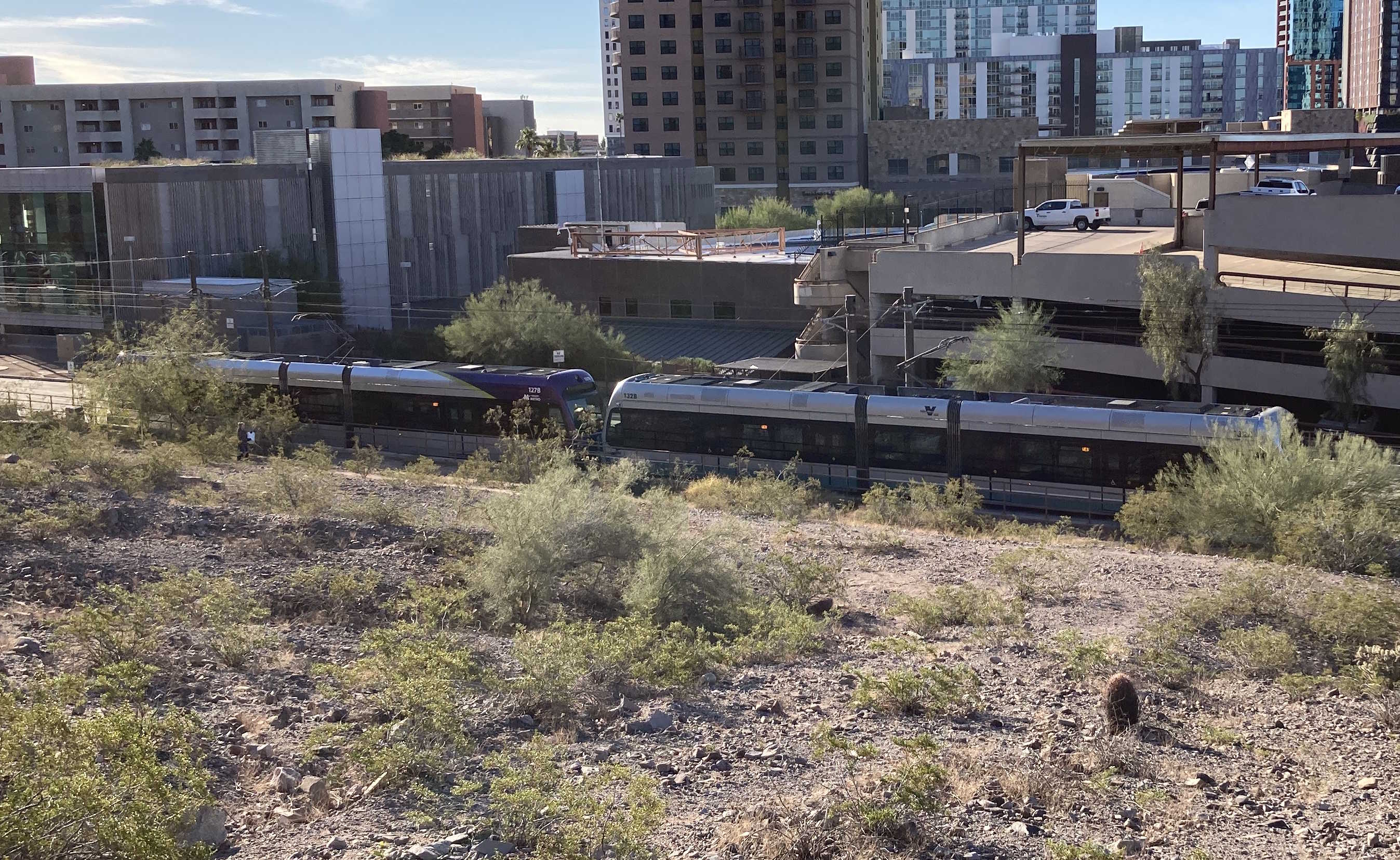 A gray and purple light rail train with a green stripe, number 127, and a silver light rail train, number 132, traveling westbound near Veterans Way and College Avenue station towards 19th Avenue and Dunlap station