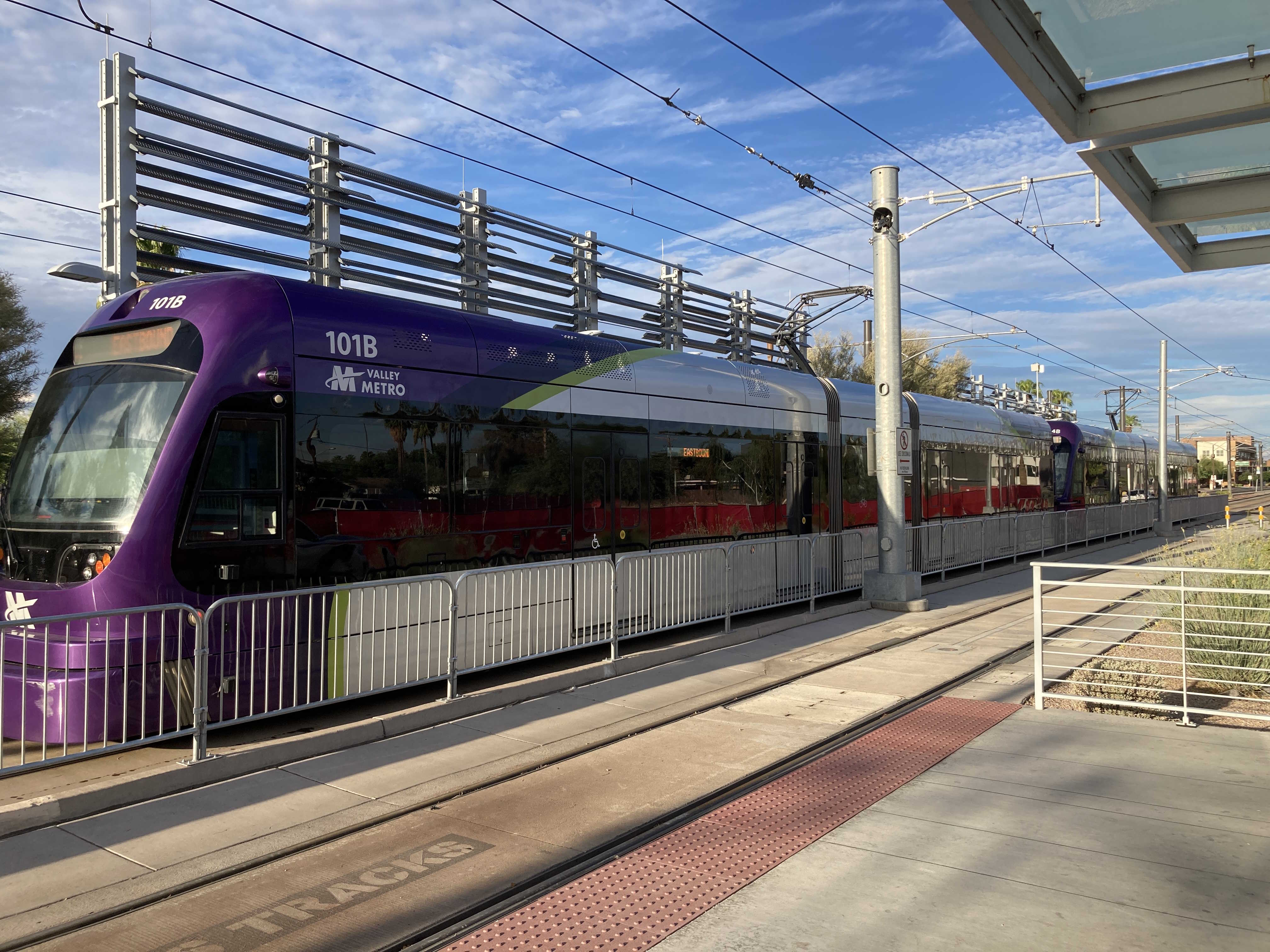 Two gray and purple light rail trains with green stripes, numbers 101 and 104, traveling eastbound on Apache Boulevard in Tempe towards Gilbert Road and Main Street station