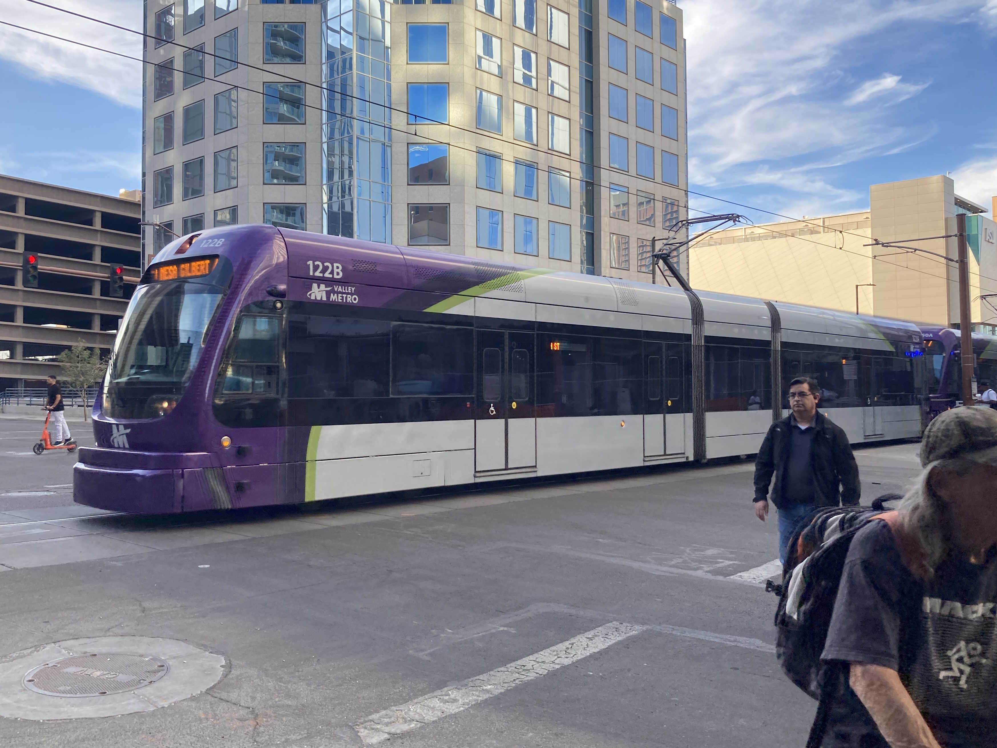 Two gray and purple light rail trains with green stripes, numbers 122 and 150, traveling southbound on First Avenue in Phoenix towards Gilbert Road and Main Street station