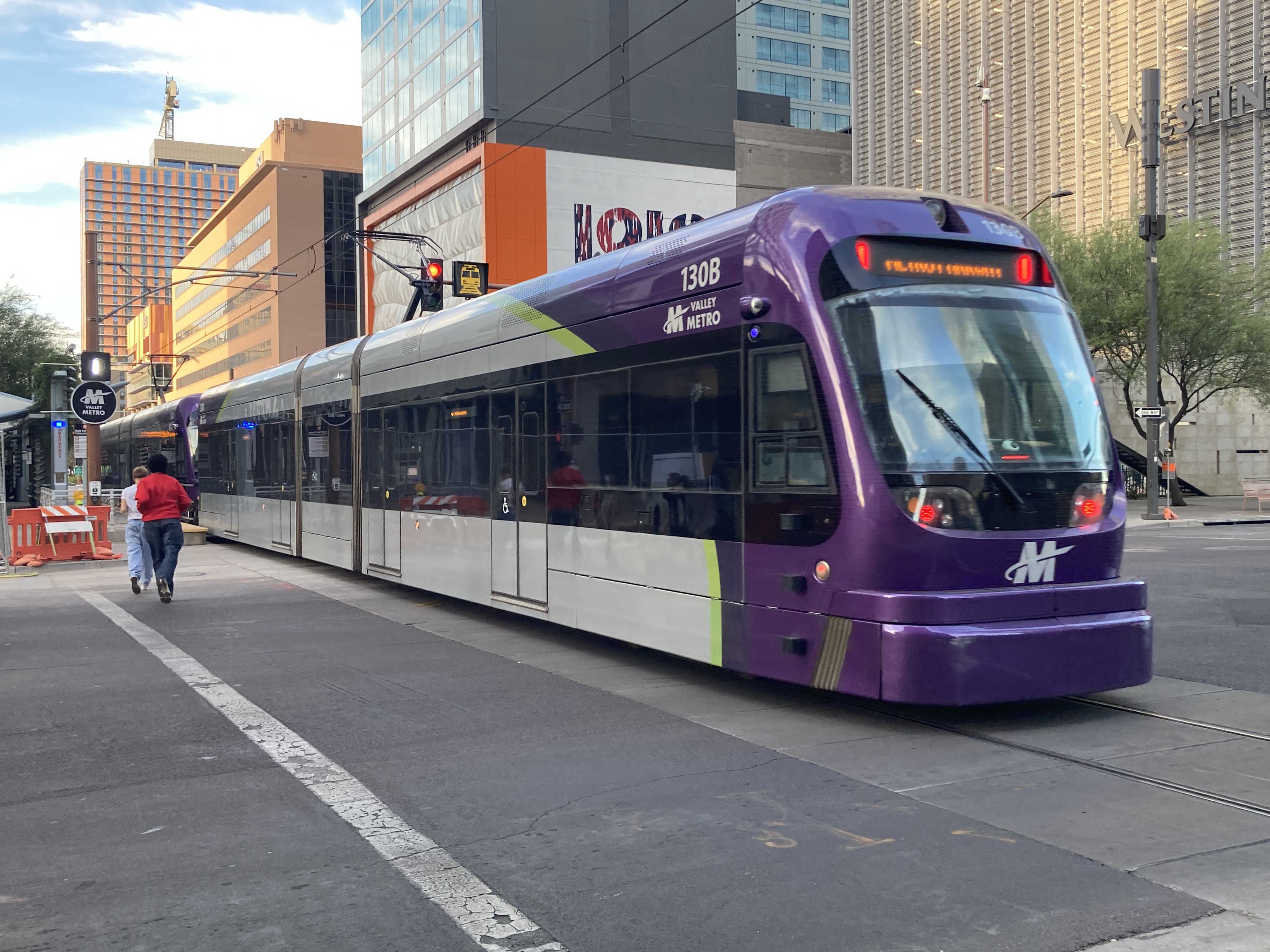 Two gray and purple light rail trains with green stripes, numbers 130 and another one unknown, traveling northbound on Central Avenue in Phoenix towards Metro Parkway station