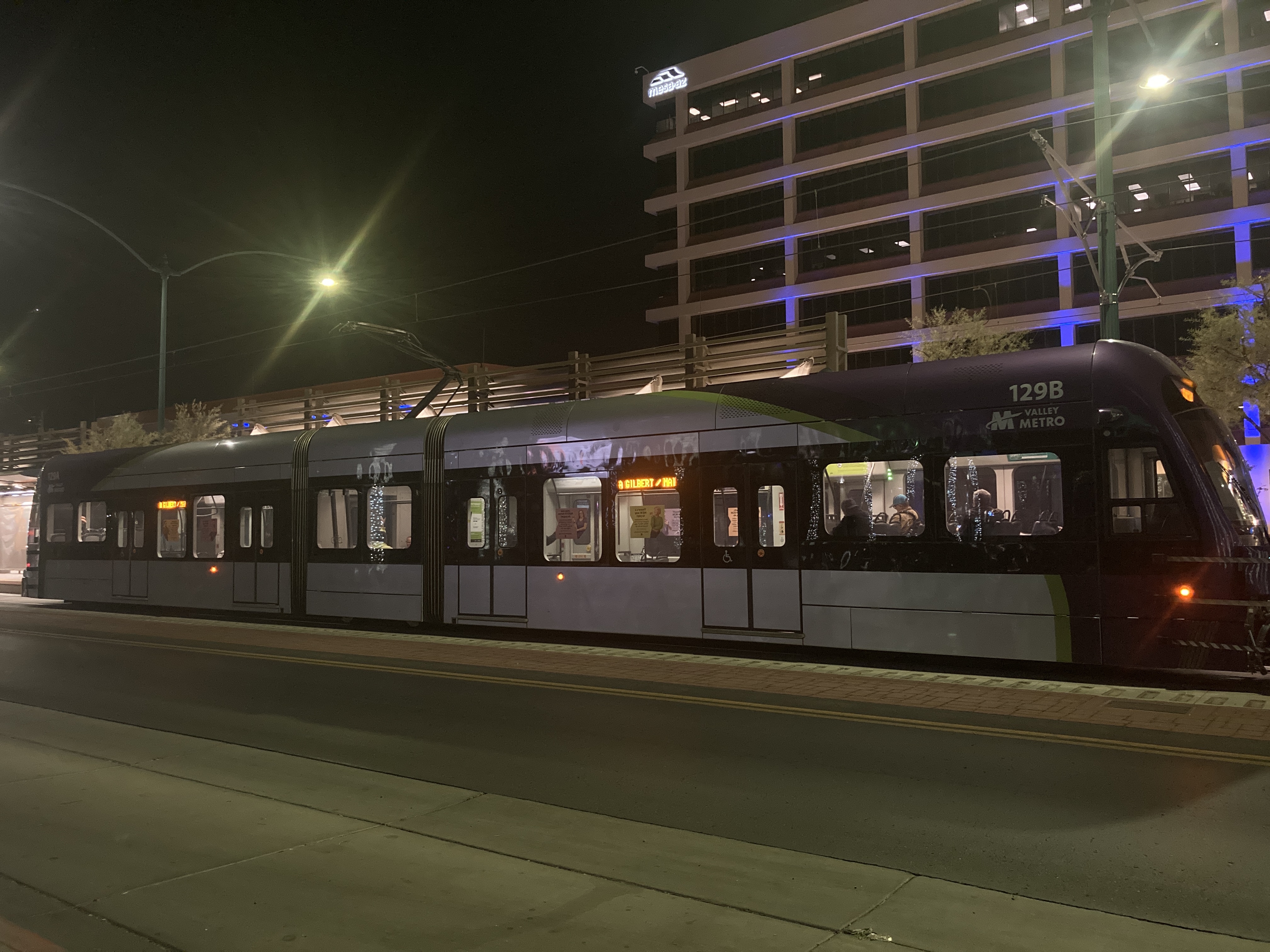 A gray and purple light rail train with a green stripe, number 129, traveling eastbound on Main Street in Mesa towards Gilbert Road and Main Street station