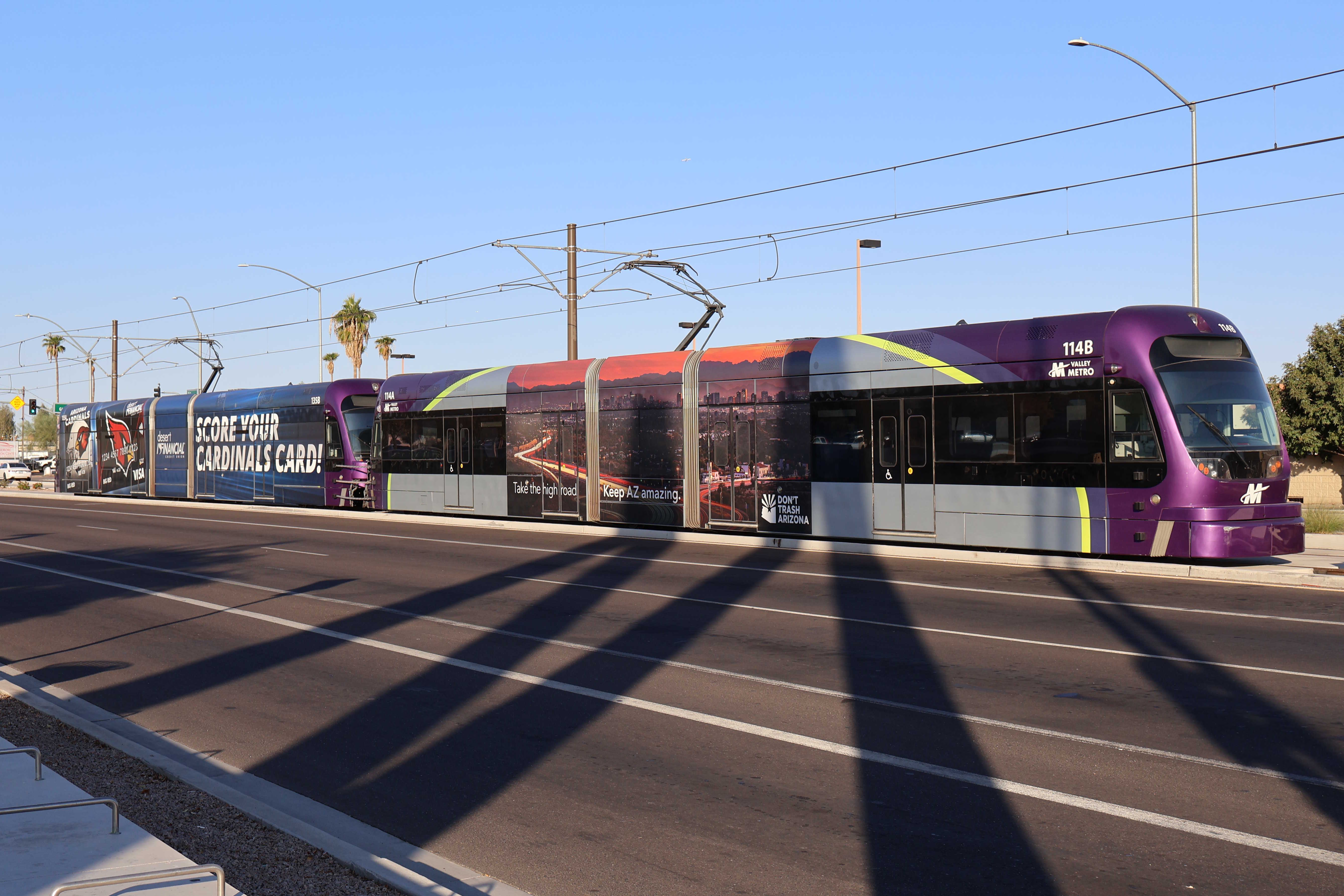 Two gray and purple light rail trains with green stripes, numbers 114 and 135, parked past the Gilbert Road and Main Street station