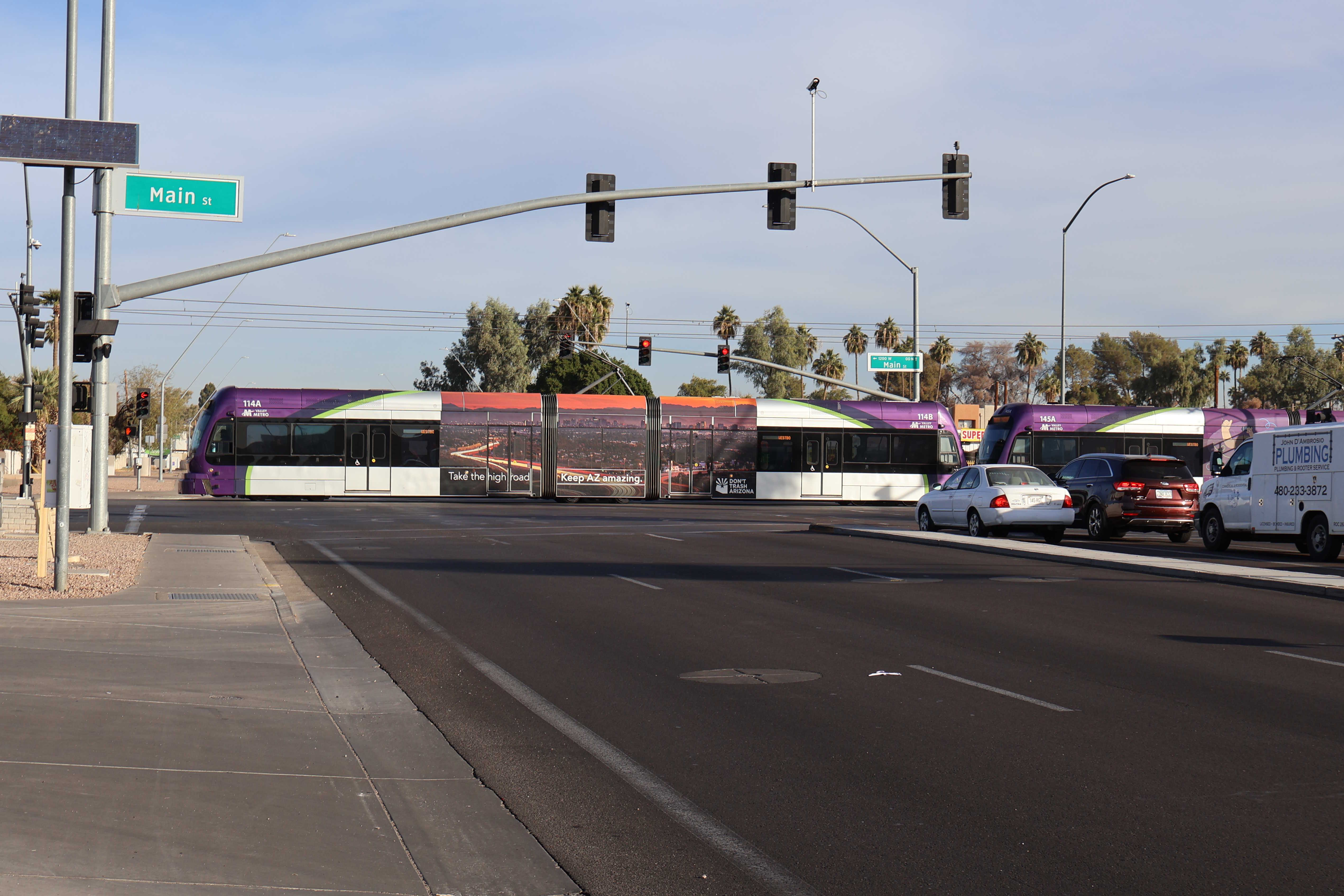 Two gray and purple light rail trains with green stripes, numbers 114 and 145, traveling westbound on Main Street in Mesa towards Metro Parkway station