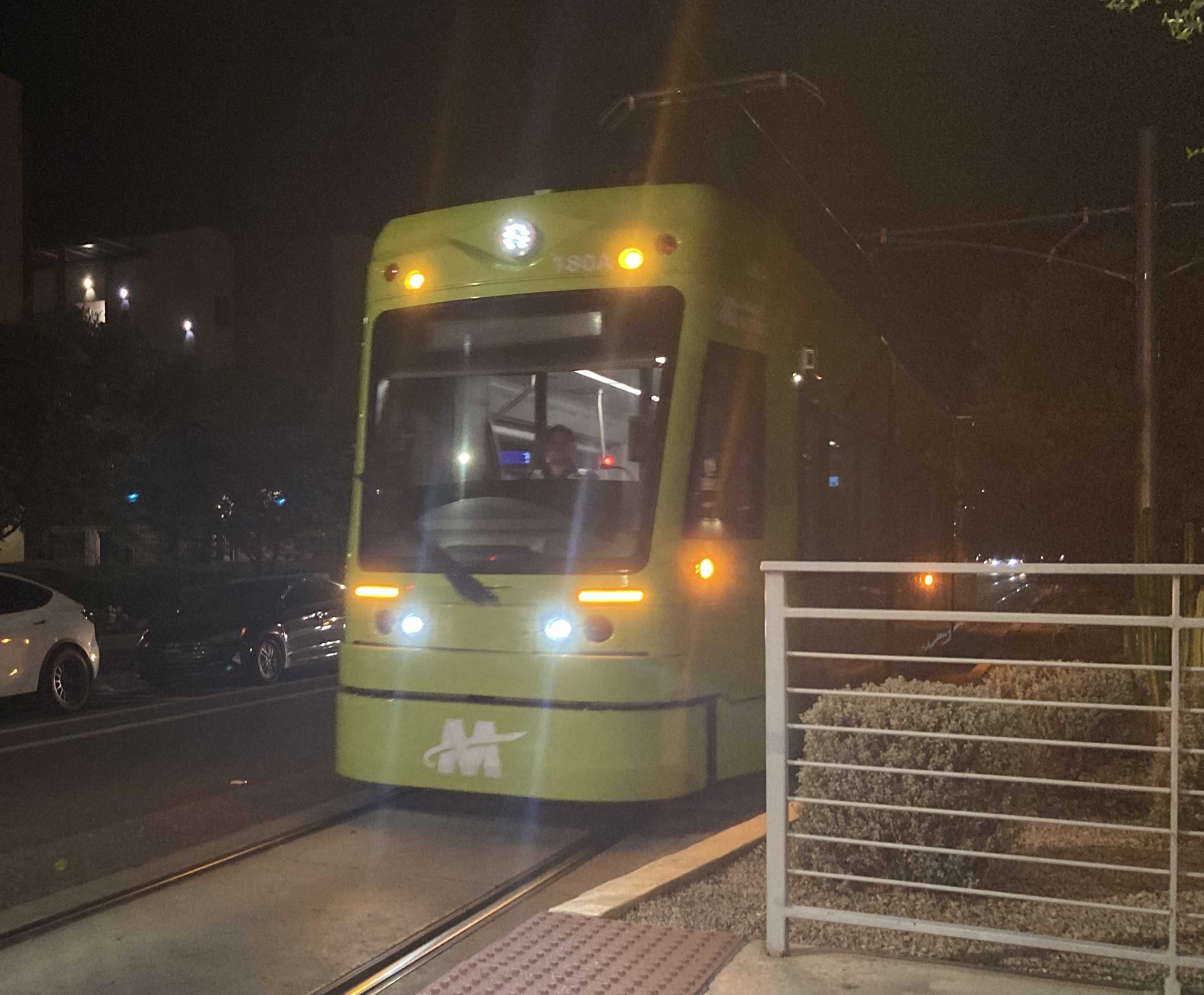 A bright green and gray streetcar with a darker green and purple stripe, number 180, at Paseo del Saber and Apache station